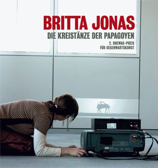 KI generiert: Das Bild zeigt eine Frau, die auf dem Boden liegt und auf einen Bildschirm schaut, der ein Bild eines tanzenden Tieres zeigt. Darüber ist der Titel "Britta Jonas - Die Kreistänze der Papageyen" und der Hinweis auf den "2. DREWAG-Preis für Gegenwartskunst" zu sehen.