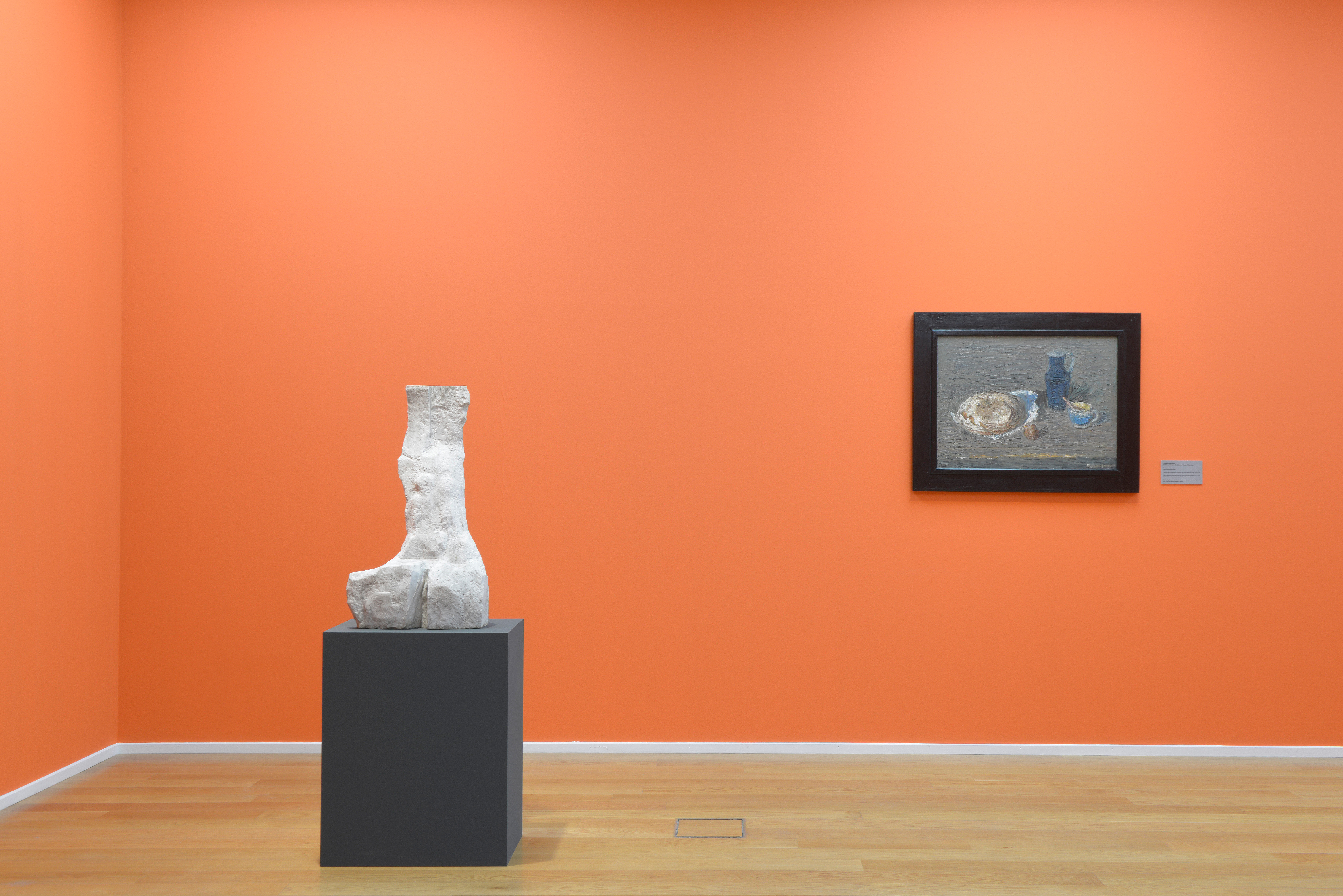 AI generated: The image shows an exhibition space with a sculpture on a plinth and a framed painting on an orange-coloured wall. The room has a wooden floor.