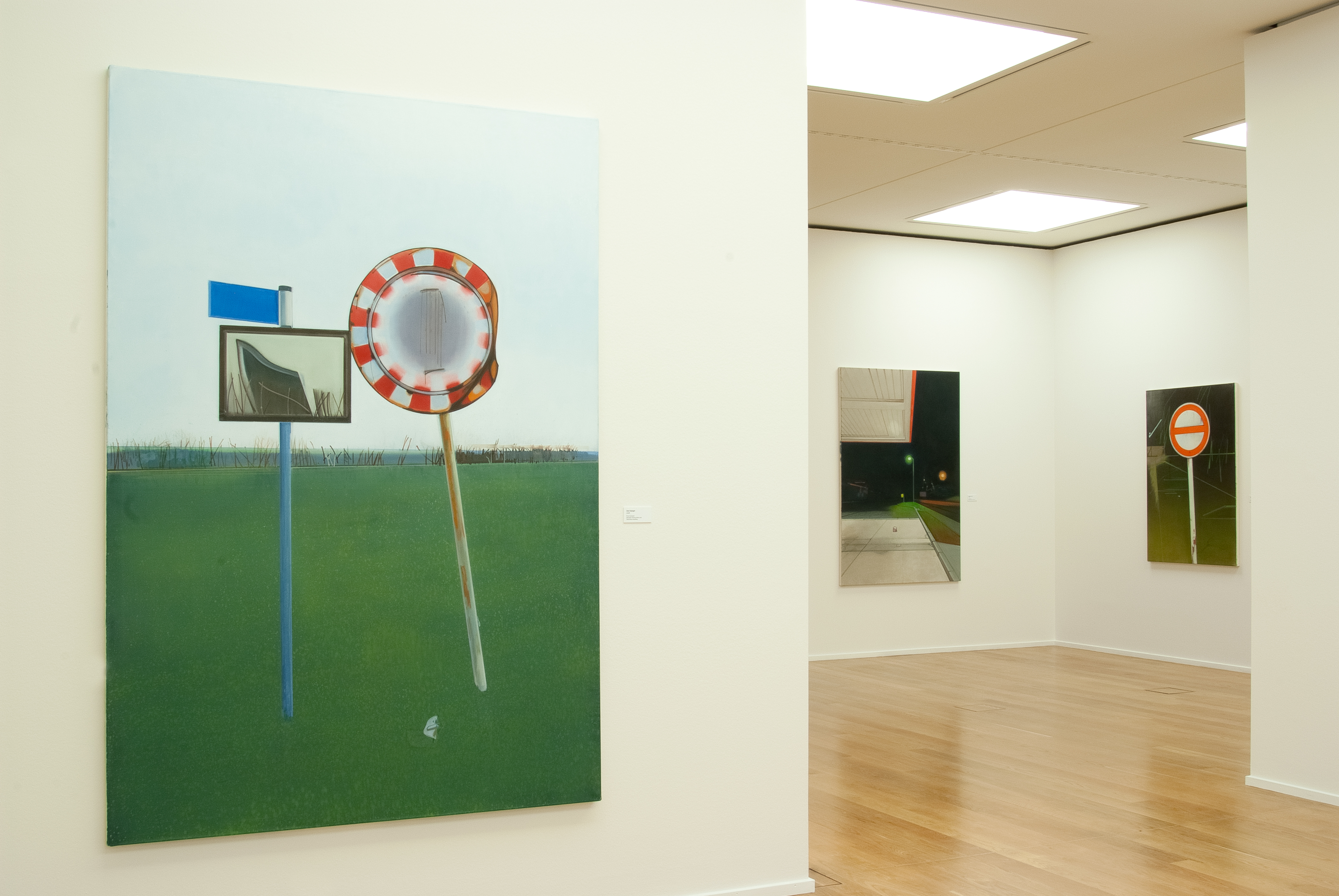 AI generated: The image shows a gallery with several large-format paintings, mainly depicting road signs and street scenes. The foreground shows a painting with a landscape theme and two eye-catching signs.