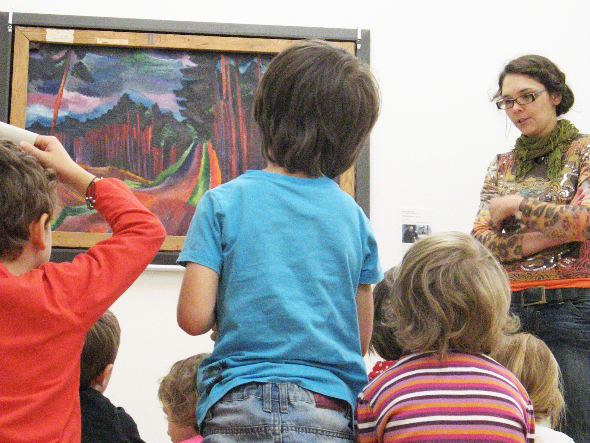 KI generiert: Das Bild zeigt eine Gruppe von Kindern, die von einer Frau vor einem farbenfrohen Gemälde in einem Museum unterrichtet werden. Die Kinder sitzen und stehen vor dem Kunstwerk, während die Frau spricht.