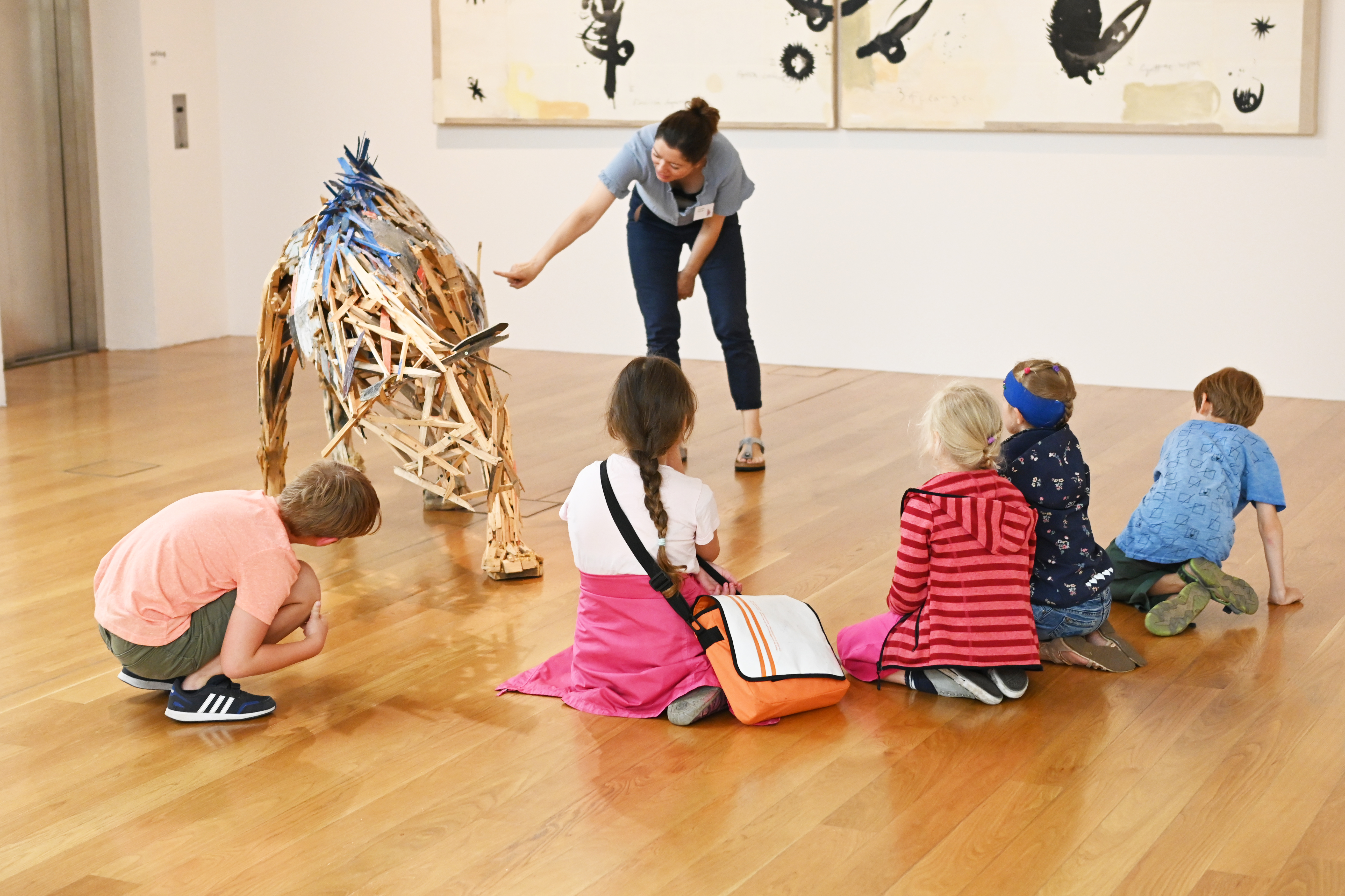 KI generiert: Das Bild zeigt eine Frau, die einer Gruppe von fünf Kindern eine kunstvolle Skulptur in Form eines Tieres erklärt. Die Kinder sitzen und stehen auf dem Boden und hören der Frau aufmerksam zu.