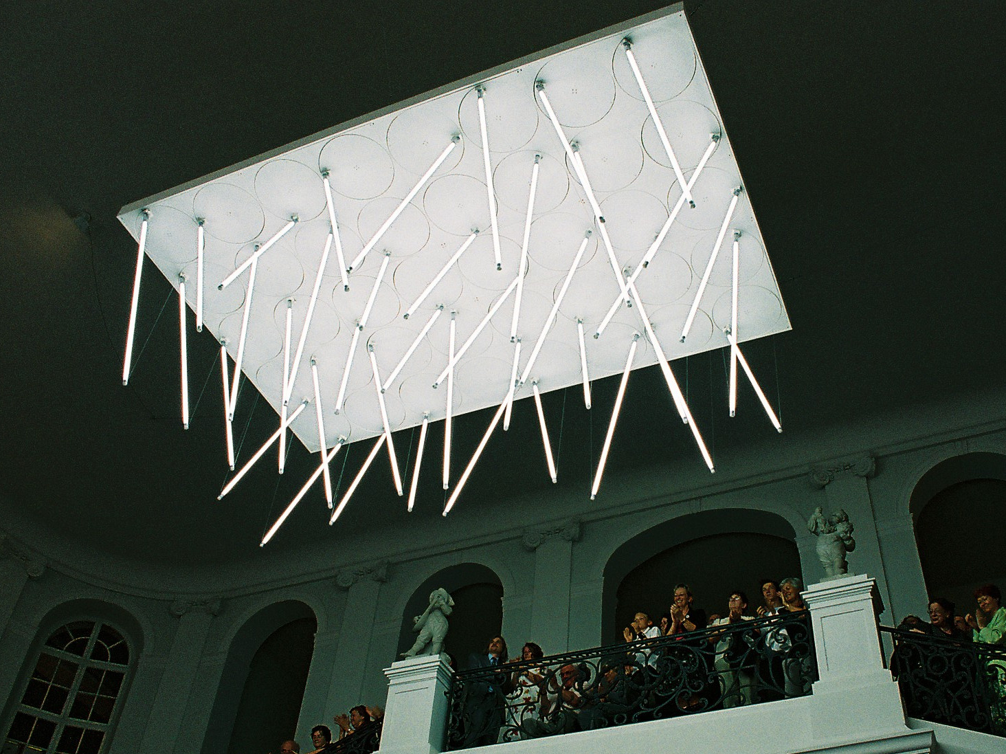 KI generiert: Das Bild zeigt eine elegante Treppenhausgalerie mit einem großen, modernen Deckenleuchter, der aus länglichen Leuchtstäben besteht. Viele Menschen stehen auf der Balustrade und schauen nach unten.