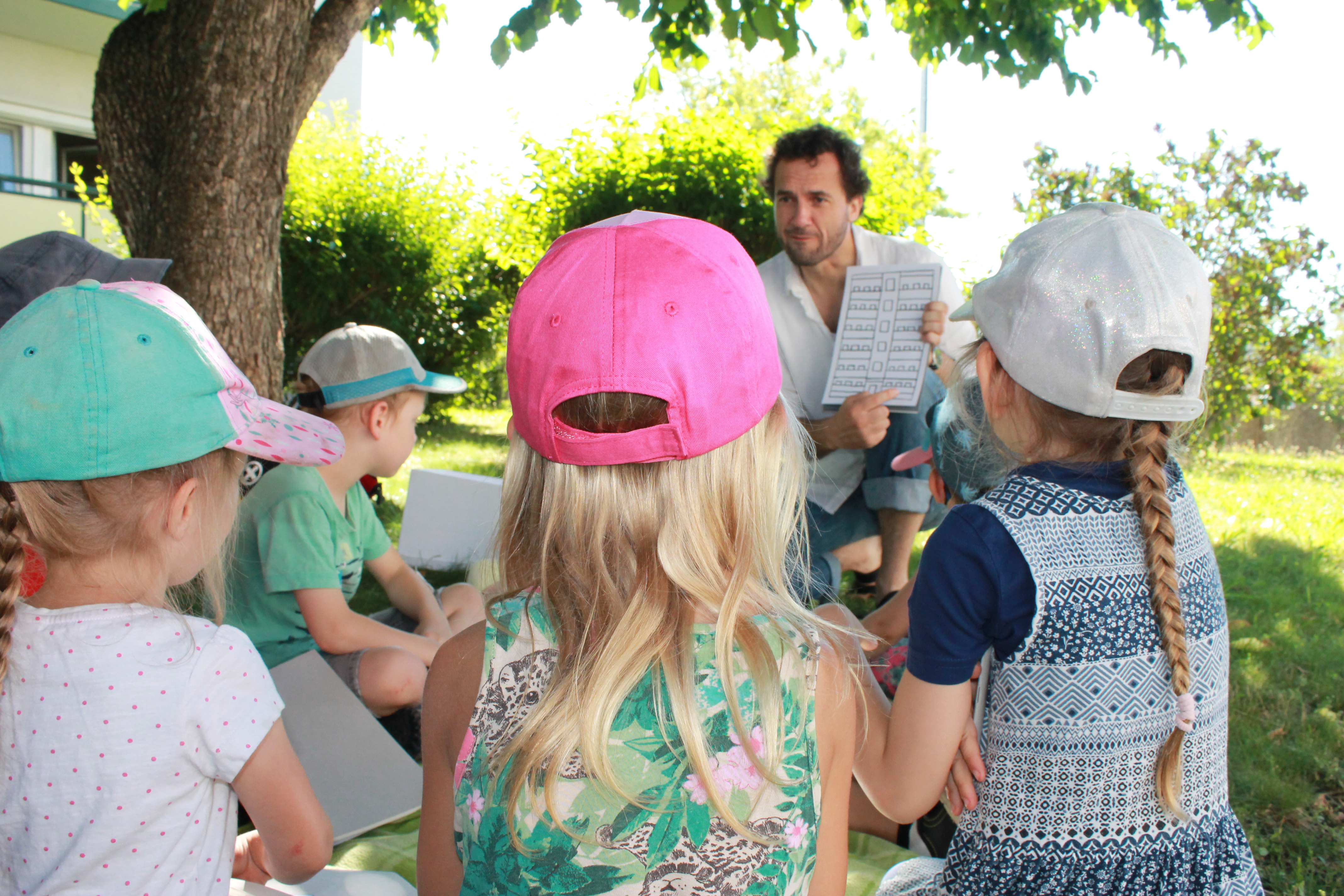 KI generiert: Das Bild zeigt eine Gruppe von Kindern, die im Freien unter einem Baum versammelt sind und einem Erwachsenen zuhören, der ihnen etwas vorliest oder zeigt. Die Kinder tragen alle bunte Baseballmützen.