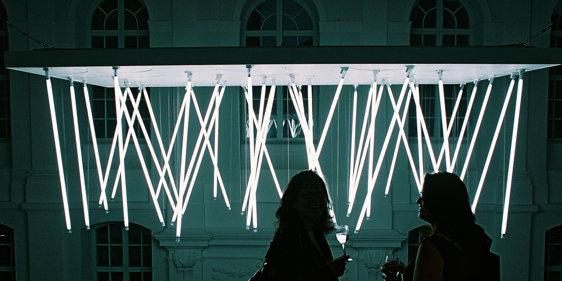 KI generiert: Das Bild zeigt zwei Personen, die vor einer beeindruckenden Lichtinstallation mit leuchtenden Röhren stehen. Der Hintergrund besteht aus einer stilvollen, balkonartigen Struktur in einem architektonischen Umfeld.