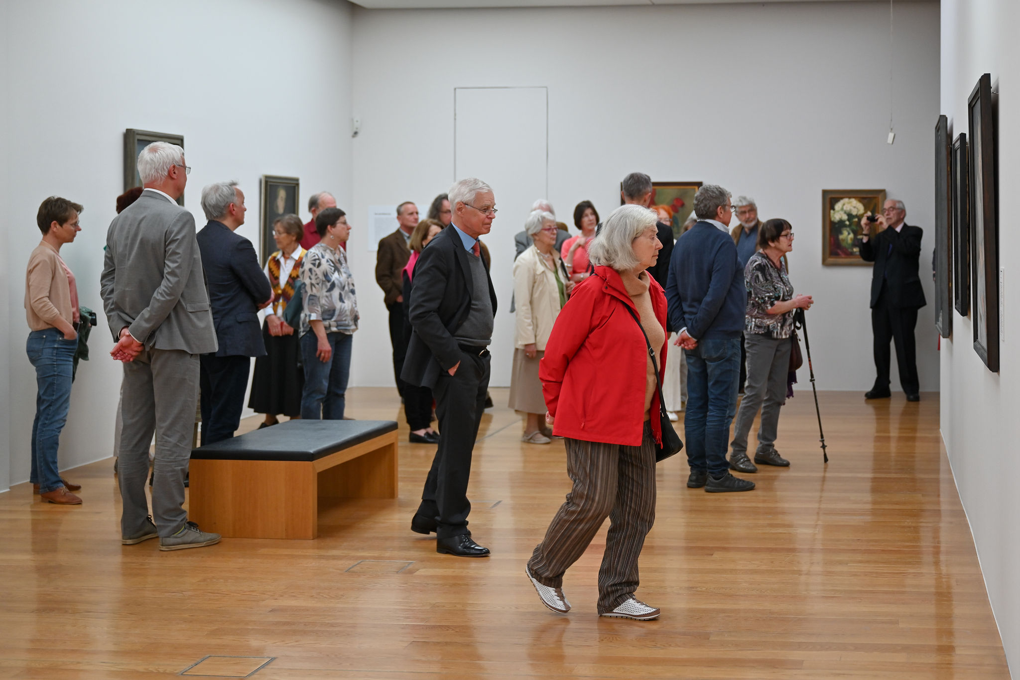 KI generiert: Das Bild zeigt eine Gruppe von Menschen, die sich in einer Kunstgalerie befinden und verschiedene Gemälde an den Wänden betrachten. Der Raum hat einen Holzboden und mehrere Kunstwerke sind ausgestellt.