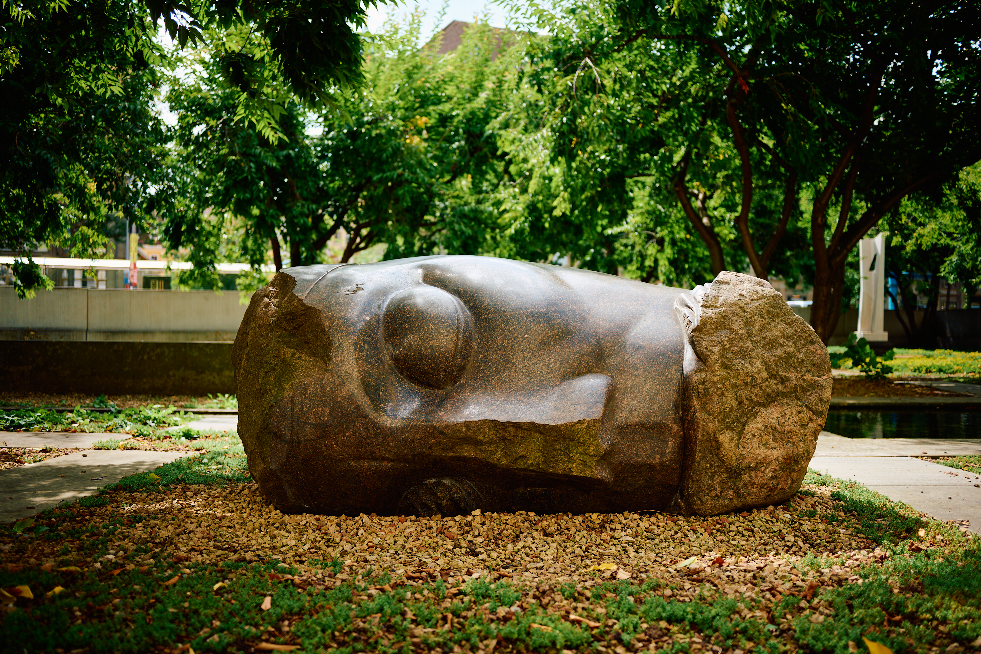 KI generiert: Das Bild zeigt eine große Steinskulptur eines liegenden Kopfes, umgeben von Bäumen und Grünflächen. Die Skulptur liegt auf einem Kiesbeet im Freien.