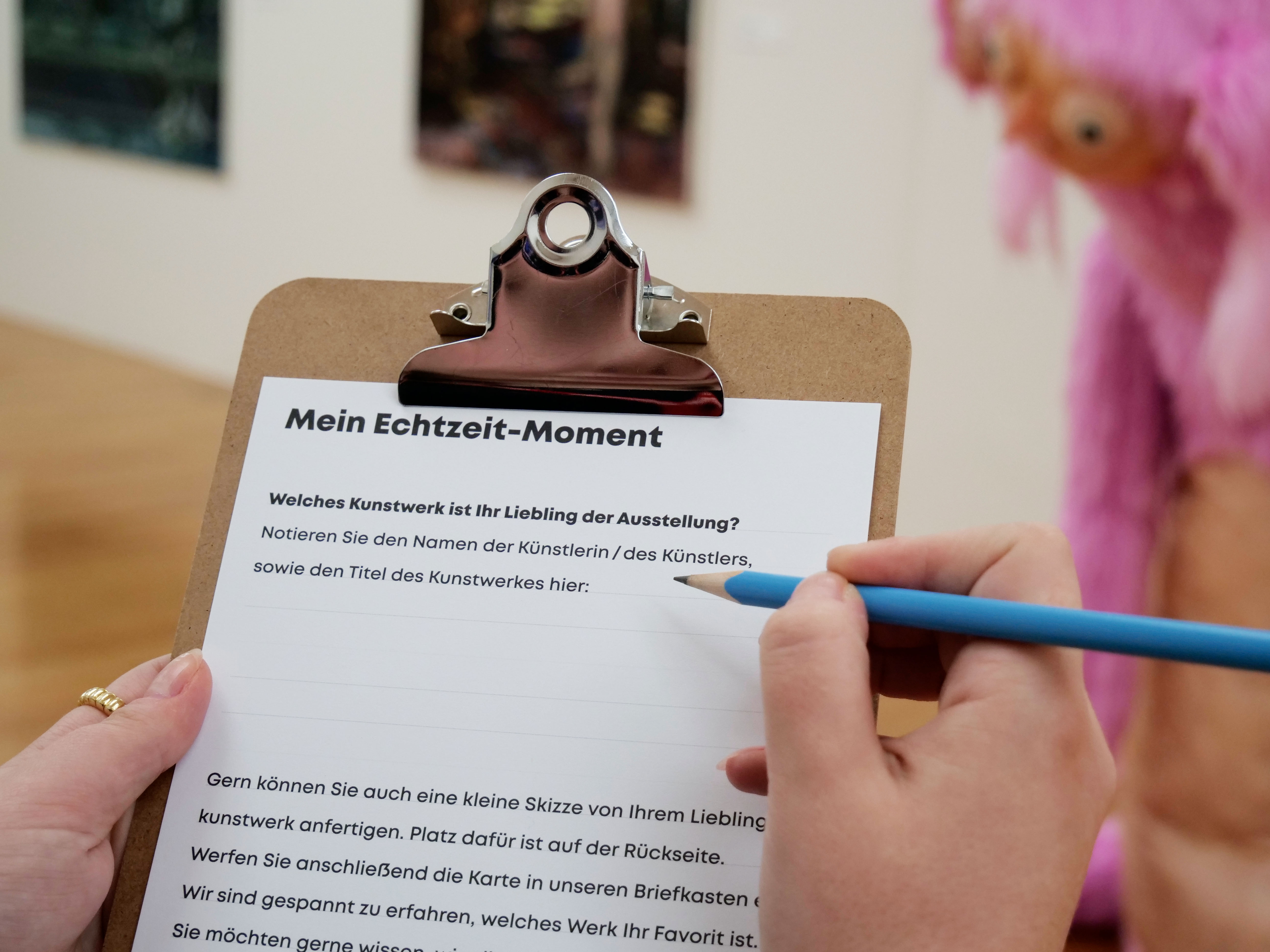 KI generiert: Das Bild zeigt eine Person, die mit einem blauen Stift auf einem Klemmbrett schreibt, welches Fragen zu Lieblingskunstwerken in einer Ausstellung stellt. Im Hintergrund ist ein unscharfes Kunstobjekt, das aussieht wie eine rosafarbene Skulptur.