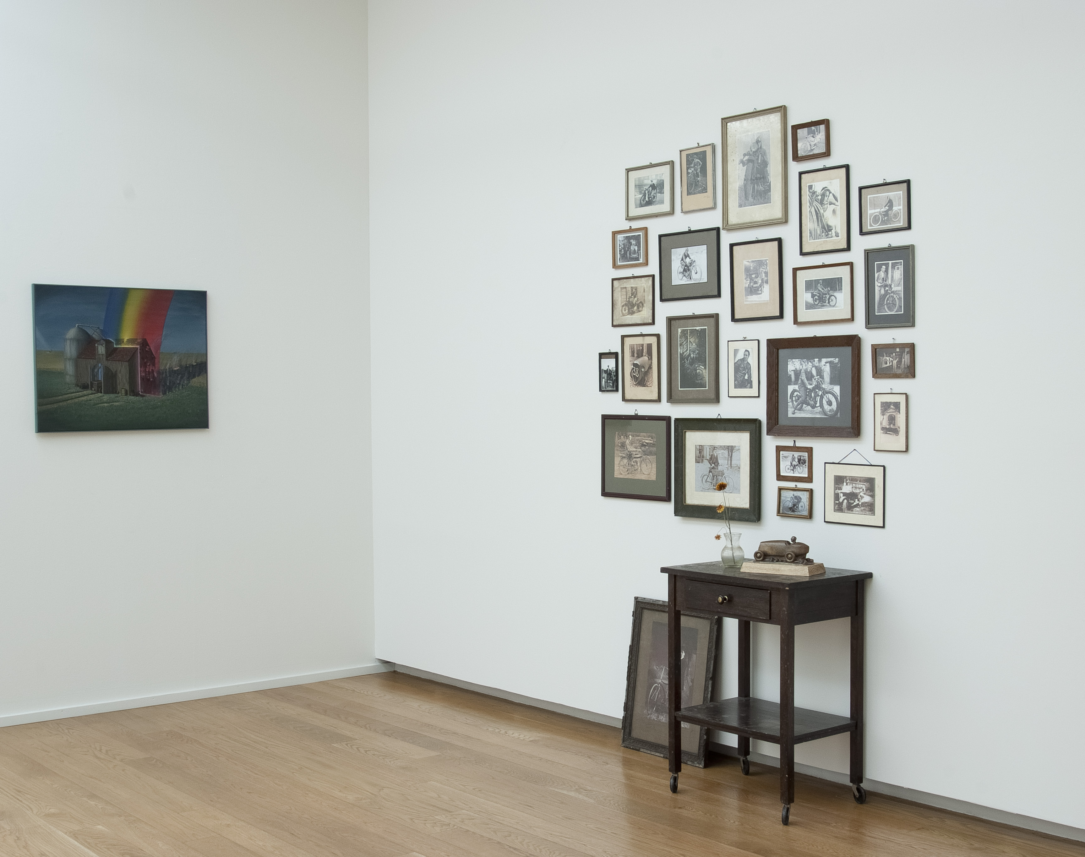 KI generiert: Das Bild zeigt eine Wand mit einer Anordnung von zahlreichen gerahmten Fotos und Drucken über einem kleinen Tisch mit ein paar Gegenständen darauf. Links an der Wand hängt ein separates Bild mit einem farbigen Regenbogenmotiv.