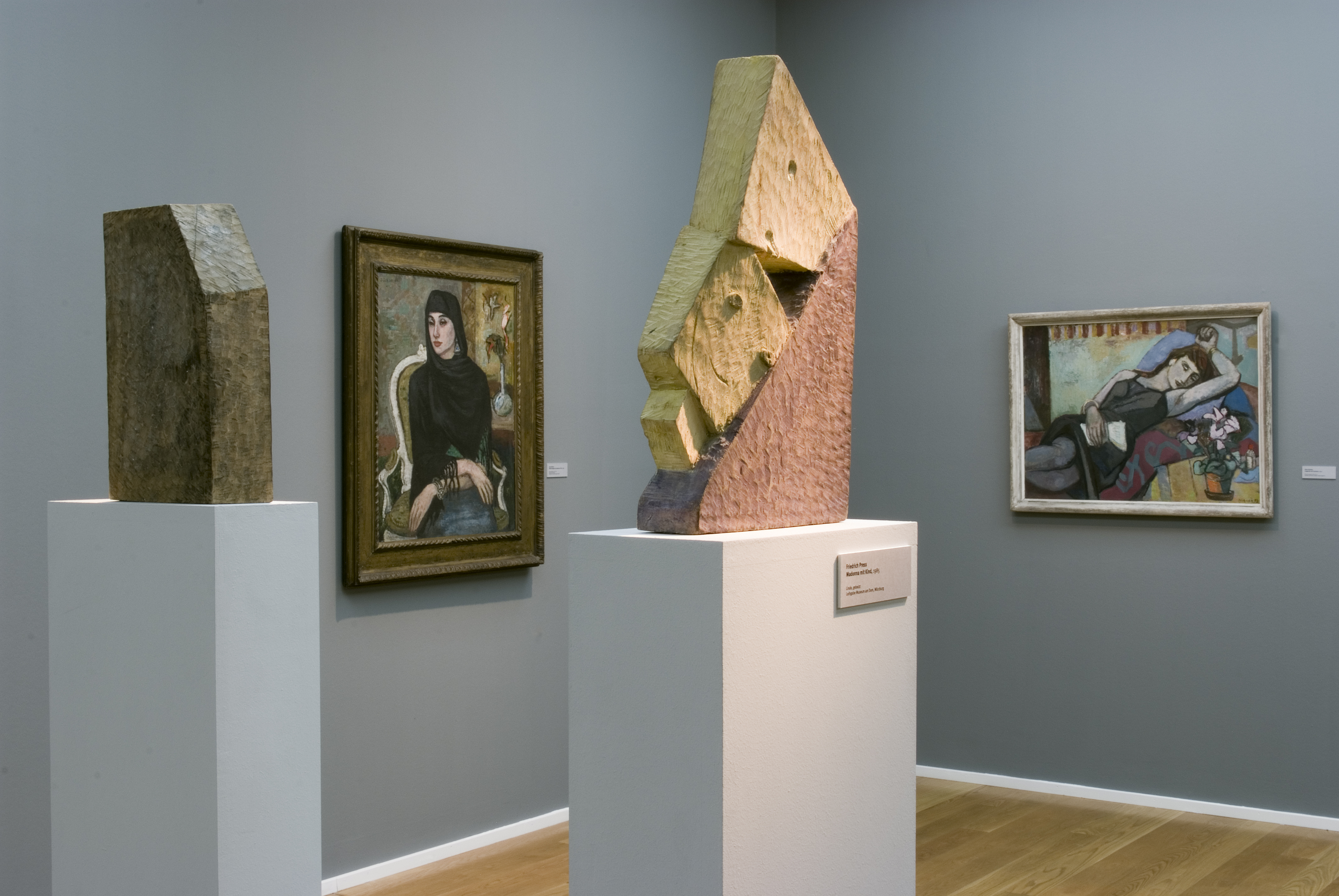 KI generiert: Das Bild zeigt eine Kunstgalerie mit zwei Skulpturen auf Sockeln und zwei Gemälden an den Wänden. Die Ausstellung kombiniert abstrakte Skulpturen mit figurativen Gemälden.