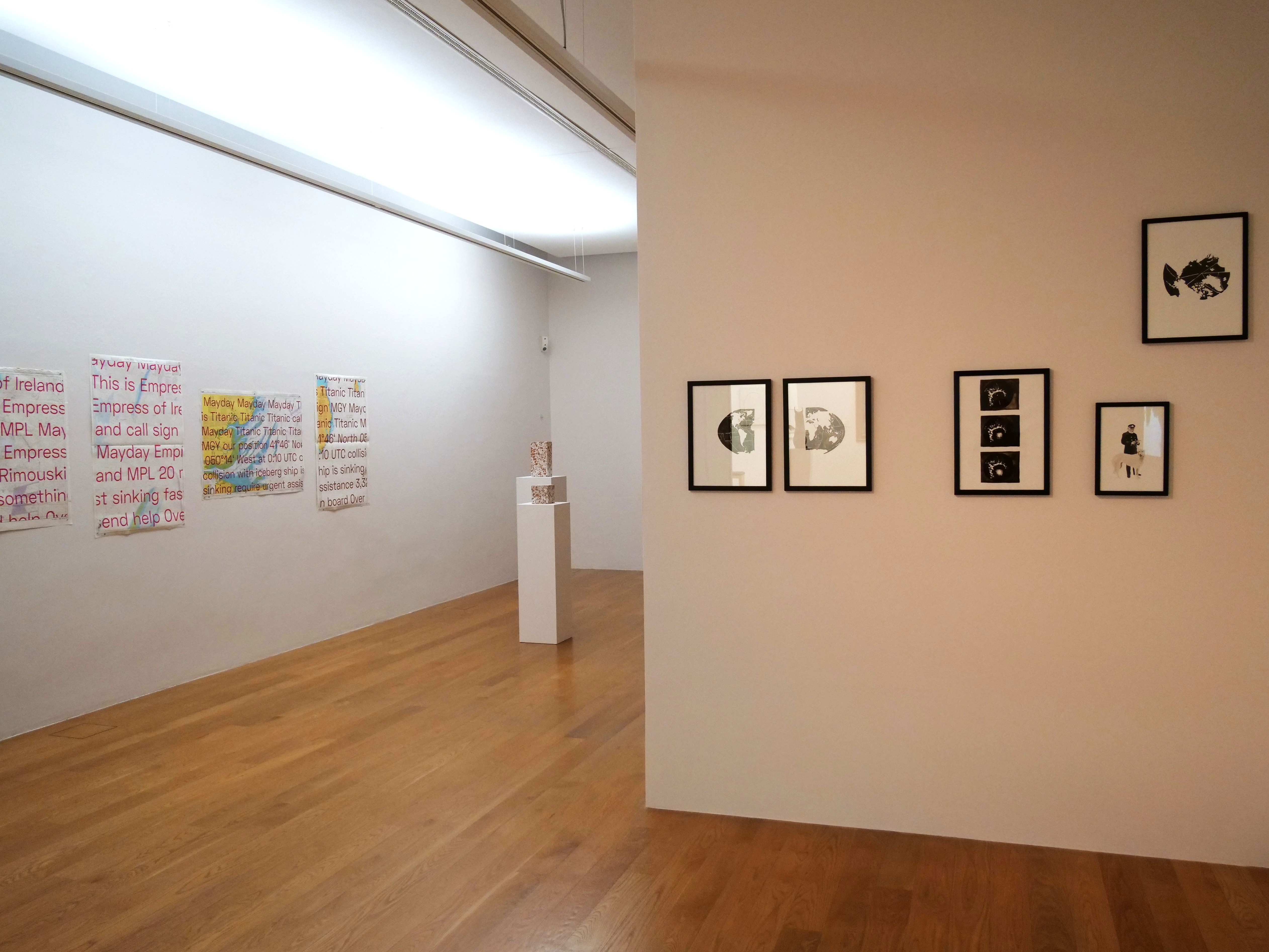 AI generated: The image shows a gallery with modern works of art hanging on white walls. There are two plinths with art objects on the floor, while large texts and black and white images can be seen on the walls.