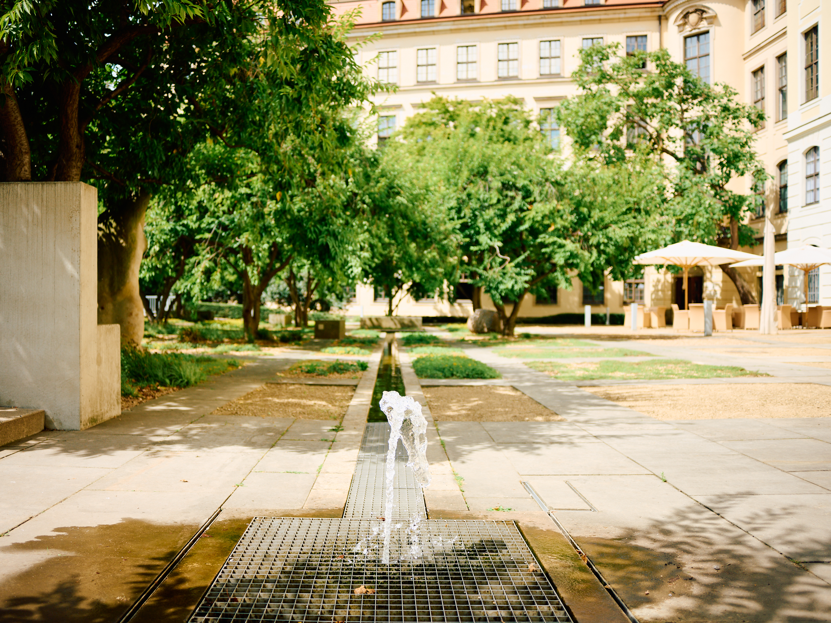 KI generiert: Das Bild zeigt einen ruhigen Innenhof mit einem kleinen Springbrunnen im Vordergrund und üppigen Bäumen im Hintergrund. Im Hintergrund ist ein historisches Gebäude zu sehen.