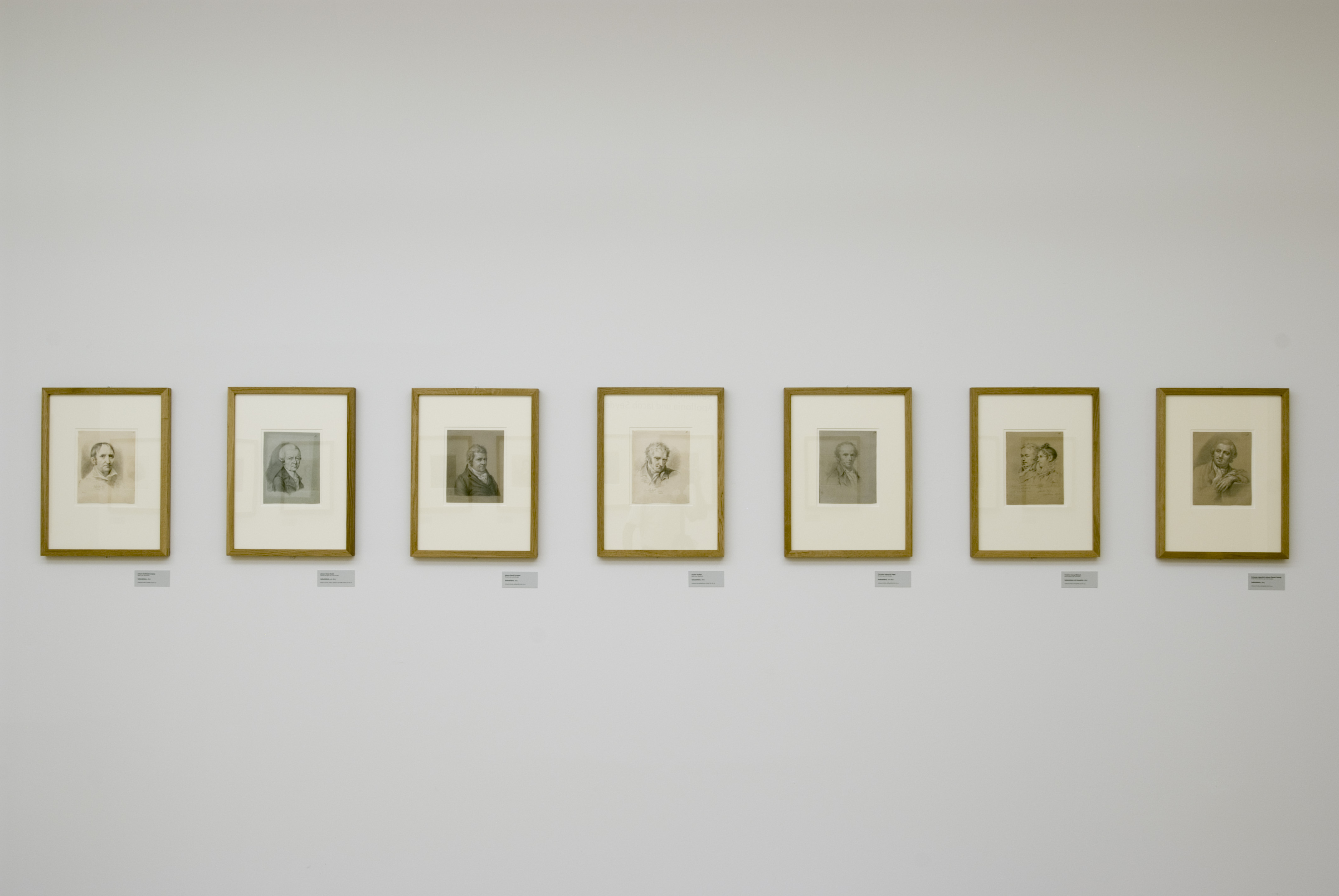 AI generated: The image shows a series of seven framed portraits hung on a white wall in a gallery. Each picture has a small caption underneath.