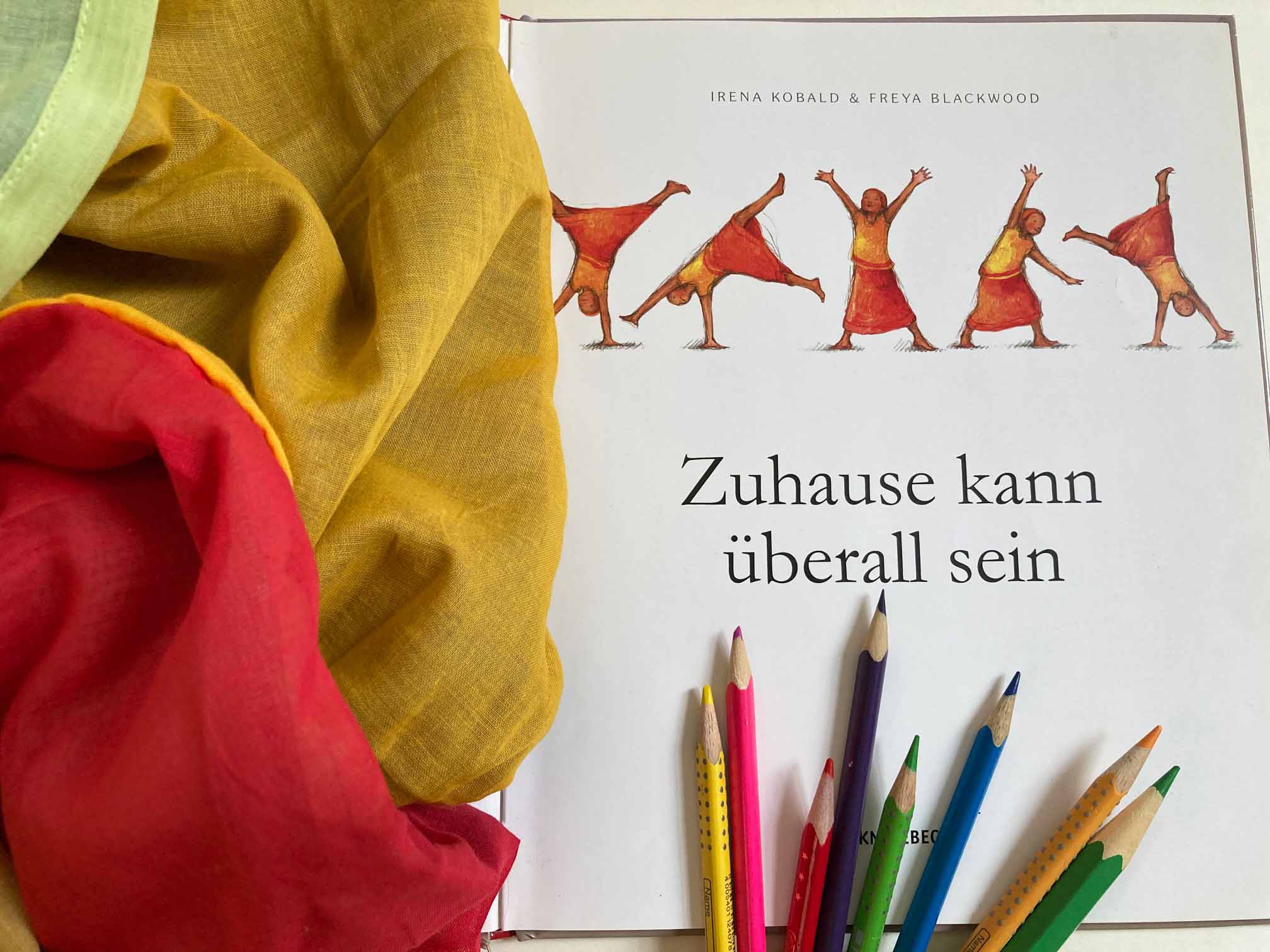 KI generiert: Das Bild zeigt ein aufgeschlagenes Buch mit dem Titel "Zuhause kann überall sein", daneben liegen Buntstifte und farbige Stoffe. Das Bild vermittelt eine kreative und farbenfrohe Atmosphäre.