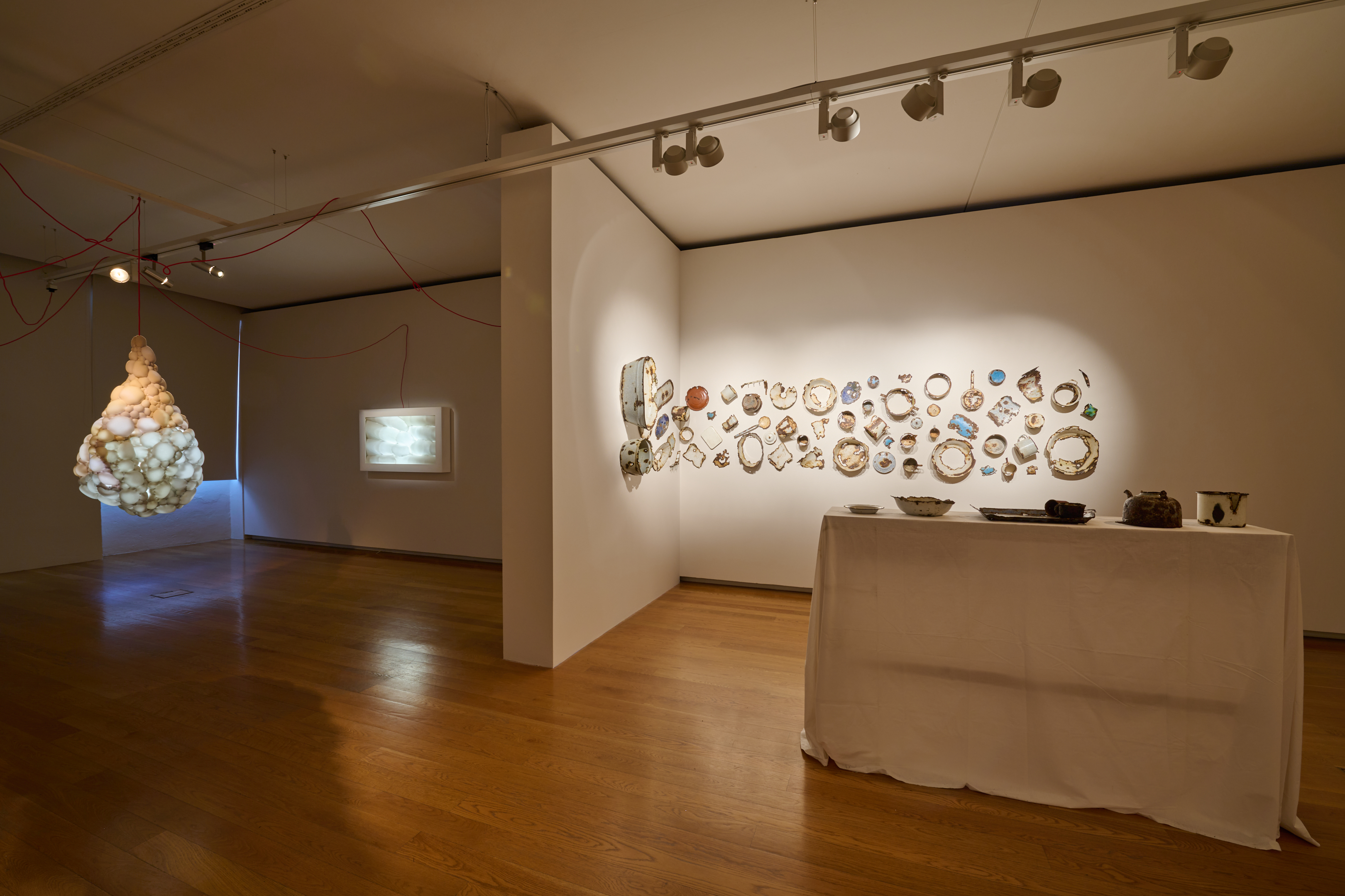 KI generiert: Das Bild zeigt eine Ausstellung in einer Galerie mit verschiedenen Kunstinstallationen. Auf der rechten Seite sind zahlreiche verzierte Teller an der Wand angebracht, während links eine leuchtende, wolkenartige Skulptur von der Decke hängt.