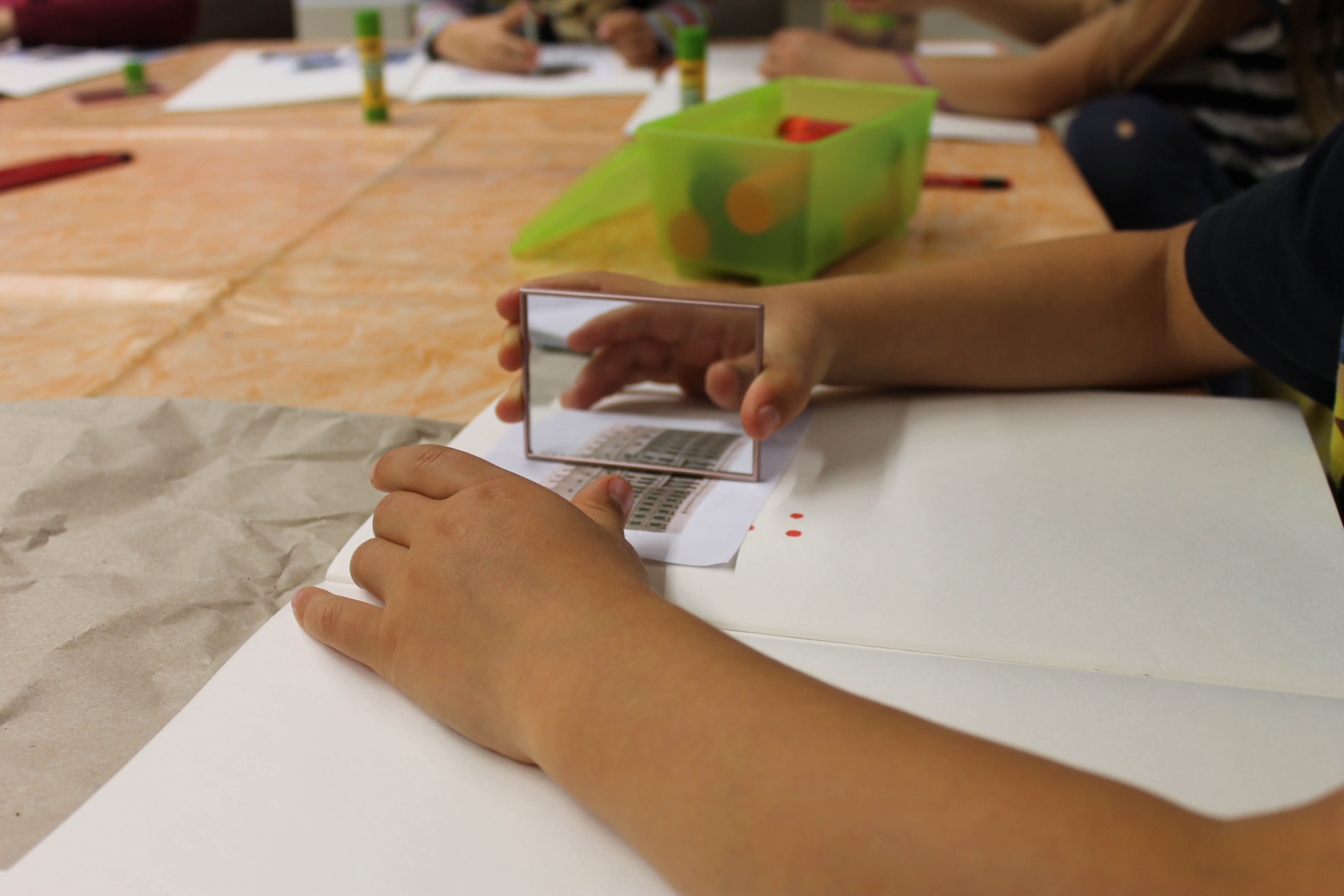 KI generiert: Das Bild zeigt ein Kind, das mit einem kleinen Spiegel auf einem Tisch arbeitet und ein Papierstück betrachtet. Im Hintergrund sind weitere Kinder zu sehen, die ebenfalls an einem Tisch sitzen und Aktivitäten durchführen.