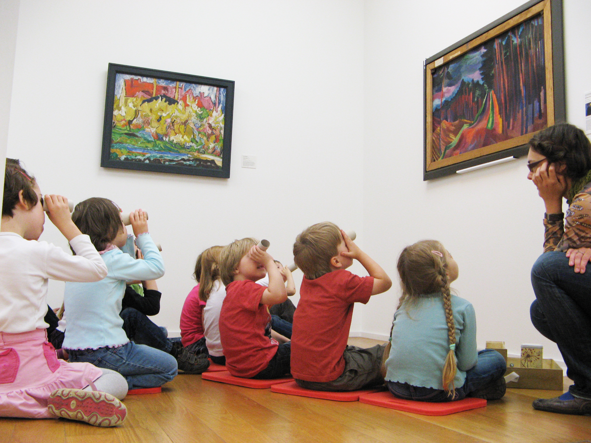 KI generiert: Das Bild zeigt eine Gruppe von Kindern, die in einem Museum auf dem Boden sitzen und Gemälde an der Wand betrachten. Eine Erwachsene führt die Kinder durch die Ausstellung und kommuniziert mit ihnen.