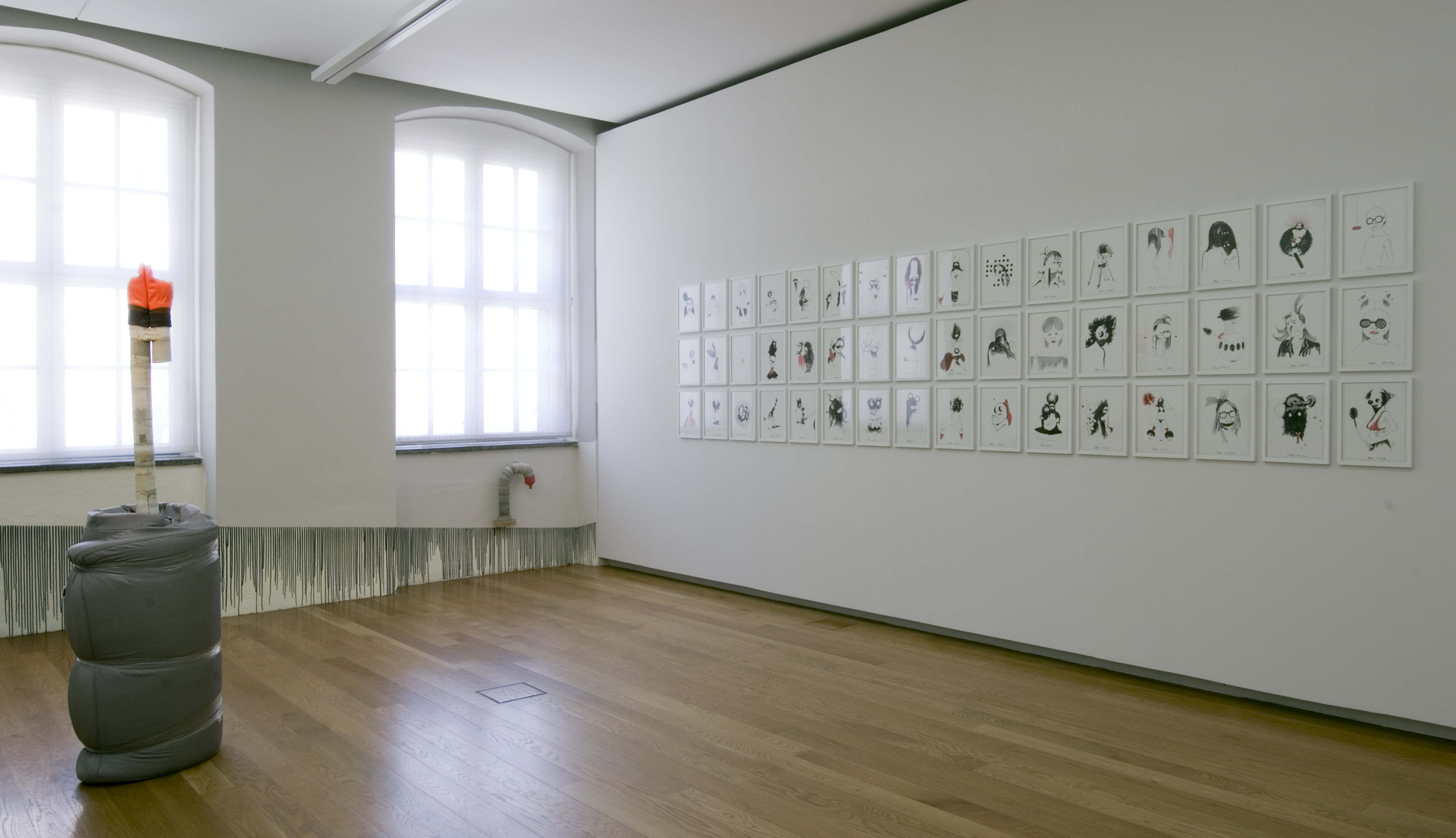 KI generiert: Das Bild zeigt einen minimalistisch gestalteten Ausstellungsraum mit einer Skulptur aus zusammengerolltem Material und einer Wand mit mehreren gerahmten Zeichnungen. Der Raum wirkt hell und modern, mit Holzboden und großen Fenstern.