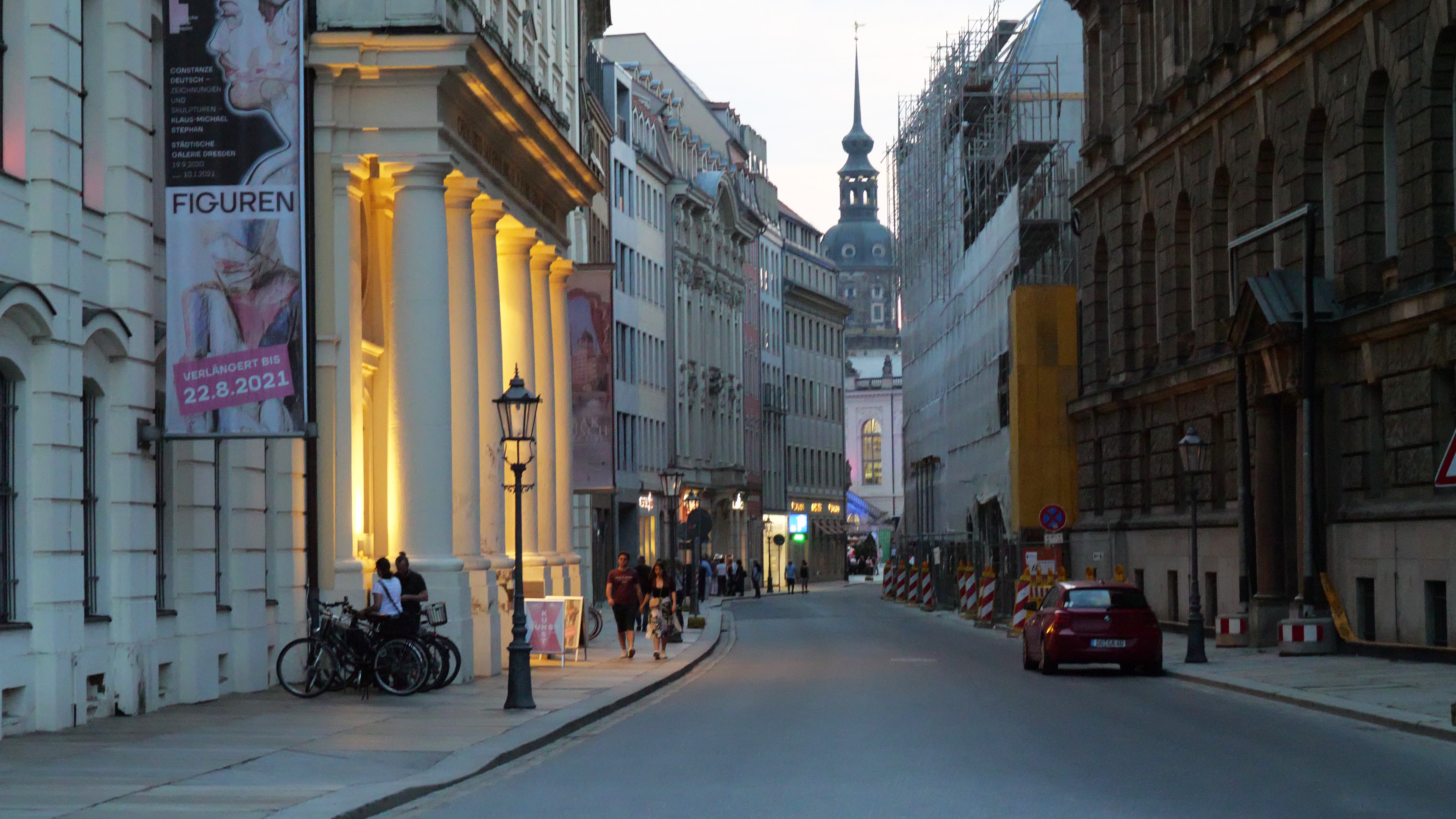 KI generiert: Das Bild zeigt eine europäische Stadtstraße bei Dämmerung, gesäumt von historischen Gebäuden und Baugerüsten. Menschen gehen an den Bürgersteigen entlang, während Autos am Straßenrand geparkt sind.