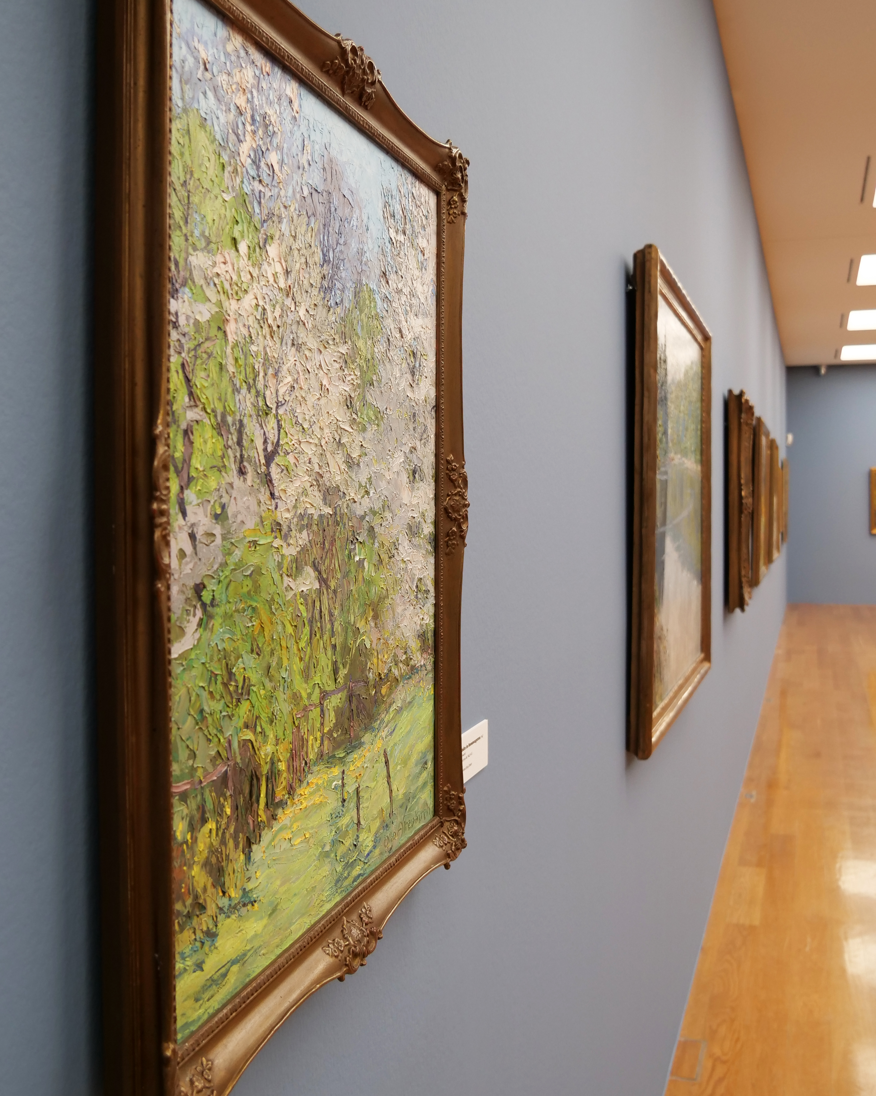 KI generiert: Das Bild zeigt eine Galleriewand in einem Museum mit mehreren Landschaftsgemälden in dekorativen Rahmen. Im Vordergrund befindet sich ein impressionistisches Gemälde mit blühenden Bäumen und grünem Laub.