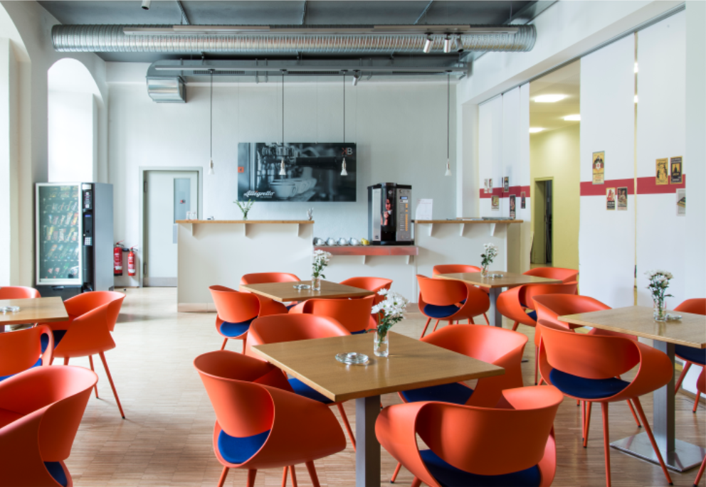 KI generiert: Das Bild zeigt eine moderne Cafeteria mit orangen Stühlen und Holztischen, an denen kleine Blumenvasen stehen. Im Hintergrund befinden sich eine Theke, eine Kaffeemaschine und ein Getränkeautomat.