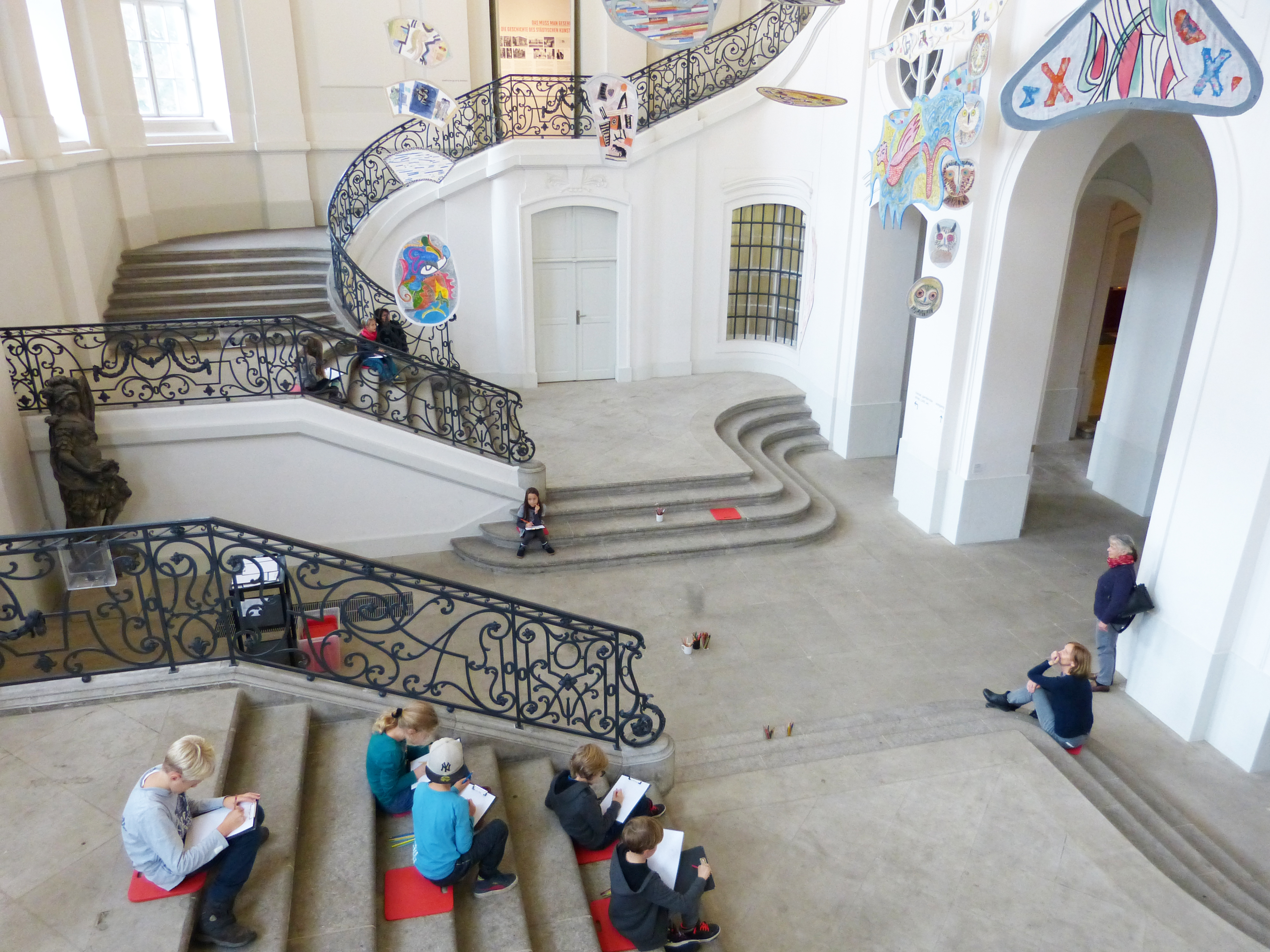 KI generiert: Das Bild zeigt eine Gruppe von Menschen, die in einem großen, lichtdurchfluteten Raum mit kunstvoll geschmückten Treppen sitzt und zeichnet. Die Hauptaktivität auf dem Bild besteht aus dem kreativen Zeichnen und sich mit dem Raum beschäftigenden Personen.