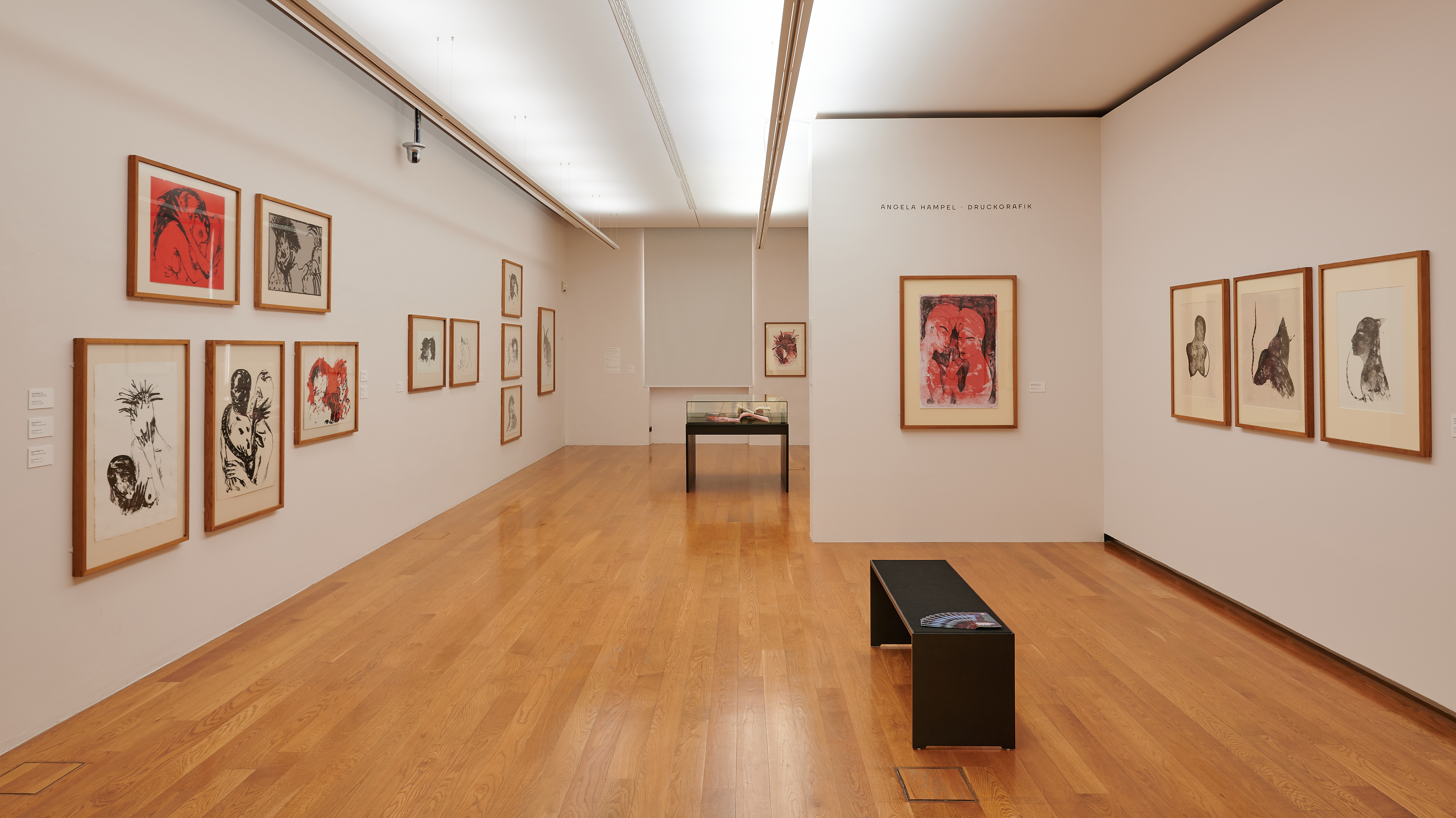 AI generated: The image shows a modern art gallery with several framed artworks on the walls, mainly comprising drawings and prints. A simple black bench stands in the centre of the room on a wooden floor.