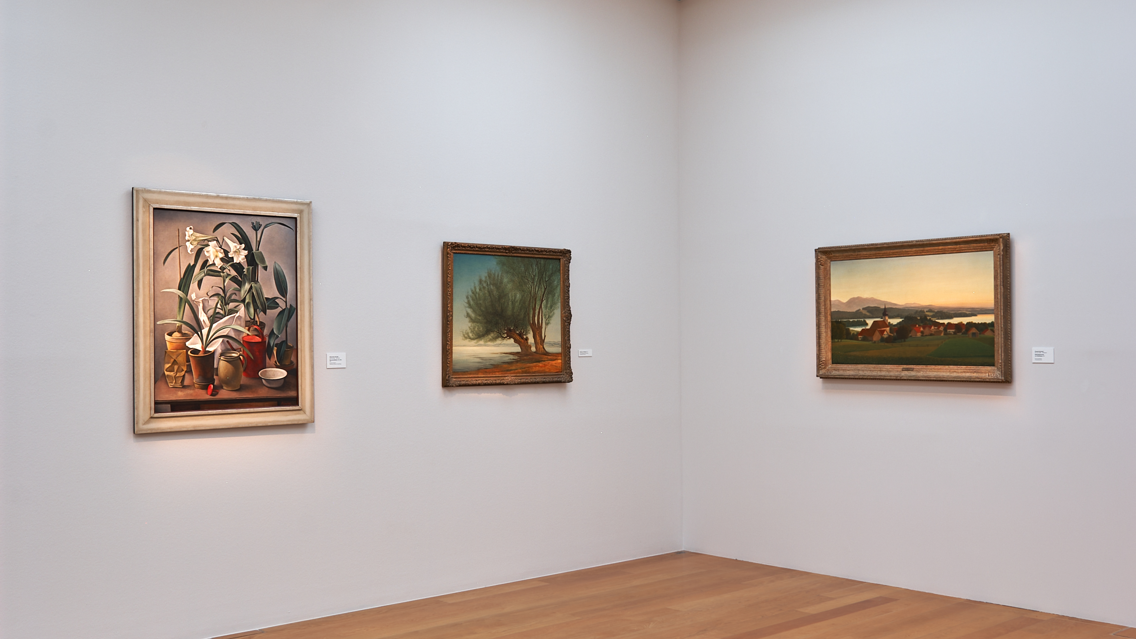 AI generated: The image shows a gallery wall with three framed paintings. The artworks show a still life, a landscape with a tree and a rural scene.
