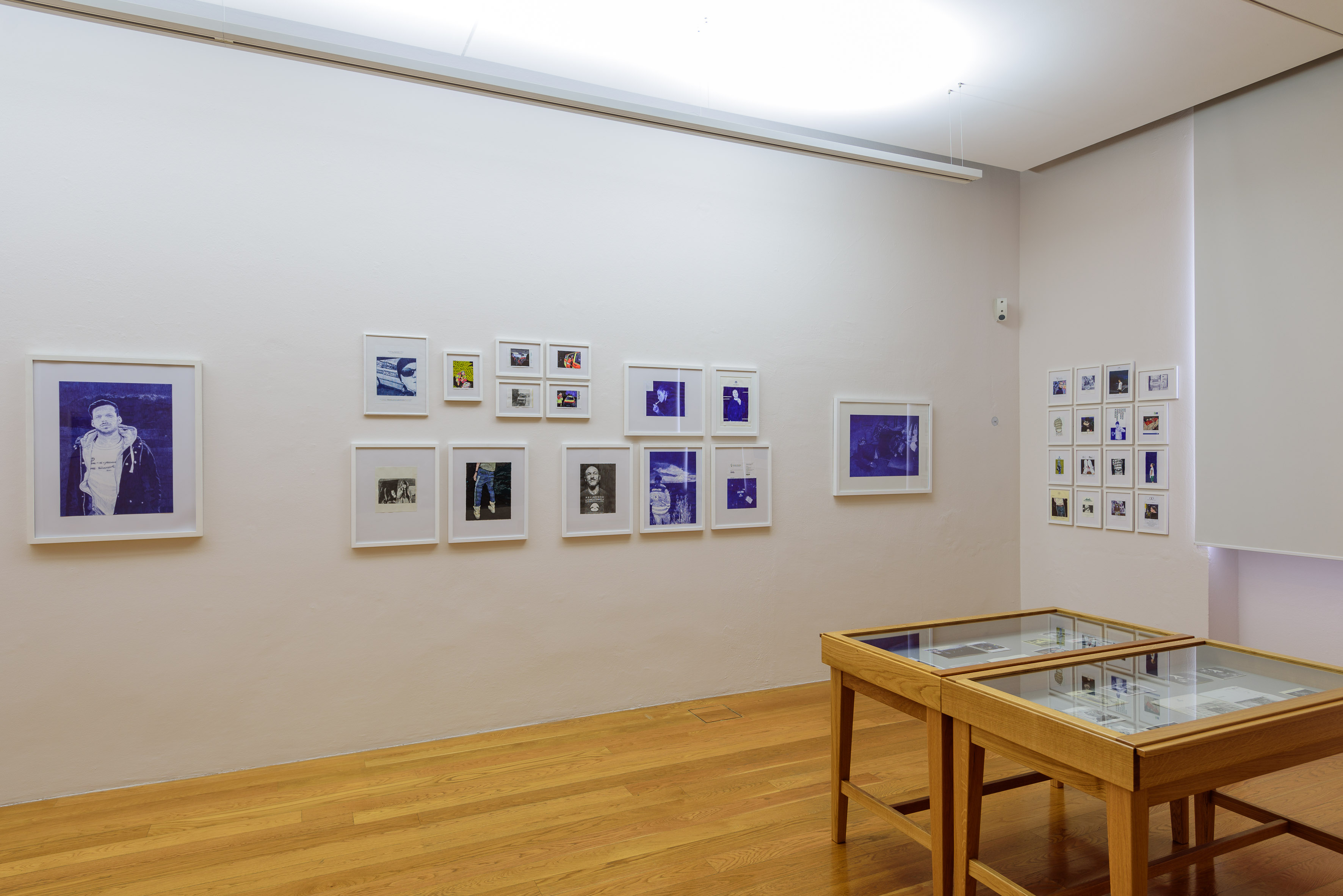 AI generated: The image shows an exhibition space with framed photographs and artworks on the walls, as well as two display cases standing on the wooden floor. The focus is on the presentation of the art and the arrangement of the pictures on the wall.