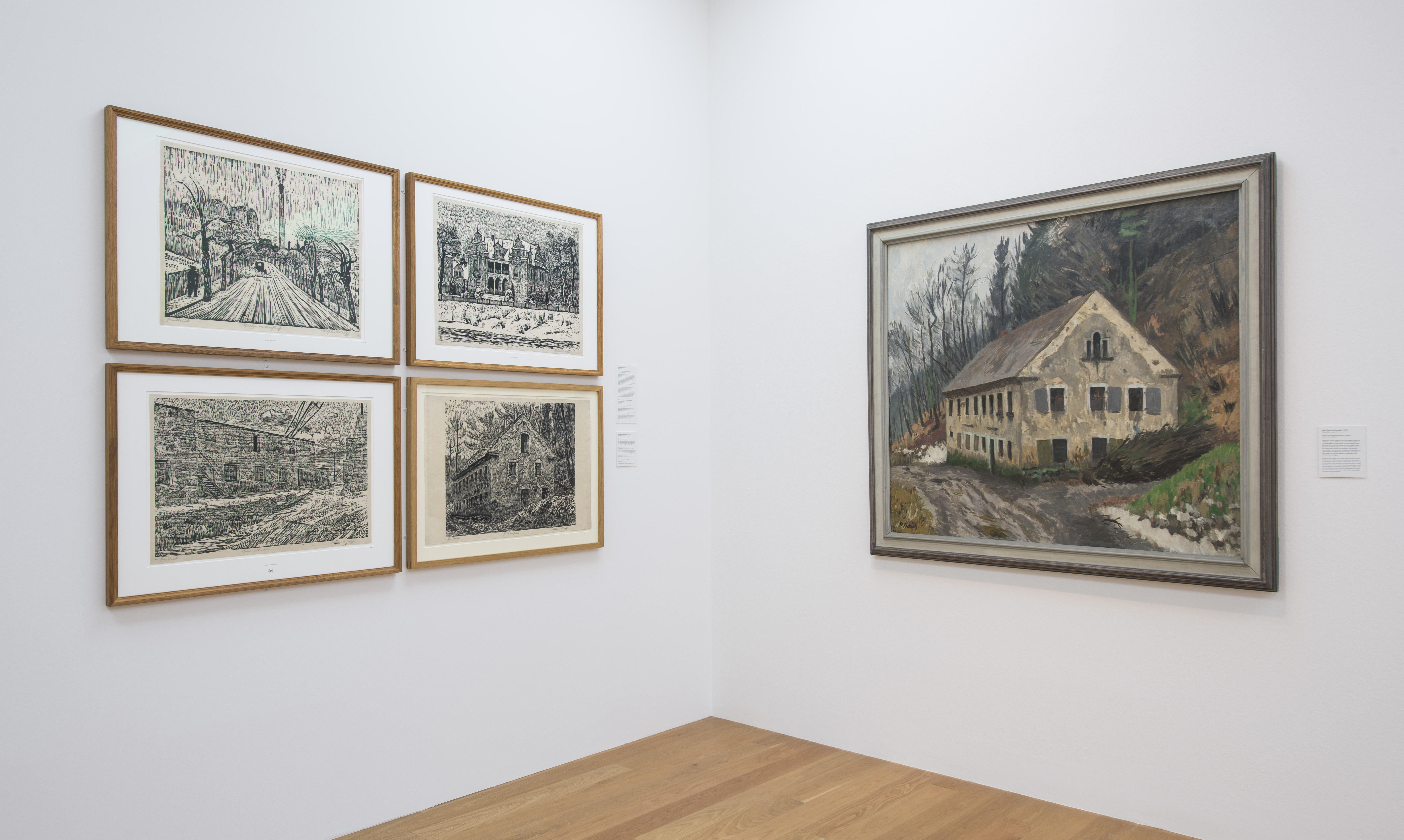 KI generiert: Das Bild zeigt eine Ausstellungswand in einem Kunstmuseum mit mehreren gerahmten Kunstwerken. Links sind vier Zeichnungen zu sehen, während rechts ein größeres Gemälde eines Hauses in einer ländlichen Umgebung hängt.