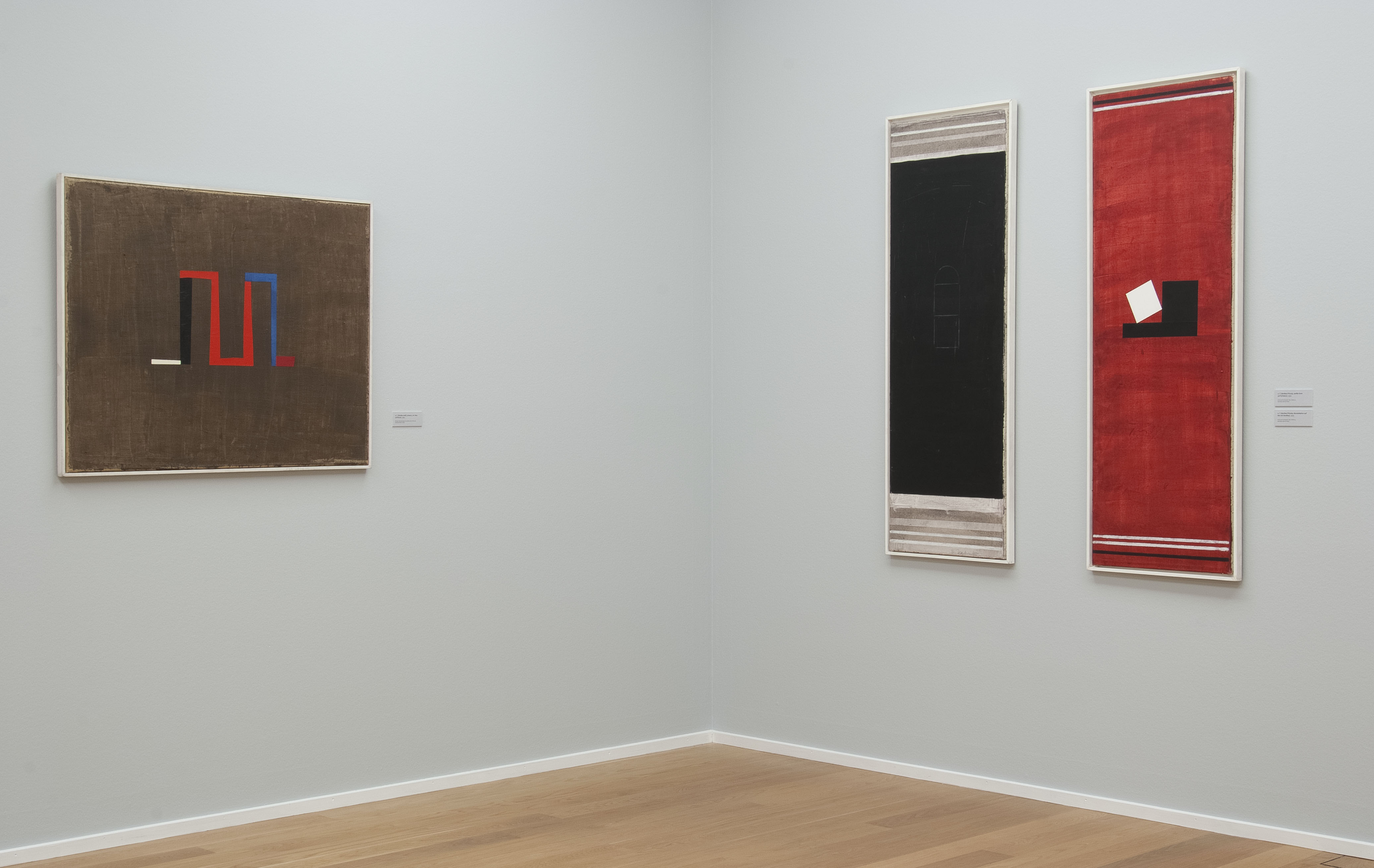 AI generated: The image shows three abstract works of art in an exhibition space, hanging on neutral walls. The works consist of geometric shapes in dominant colours such as red, blue and black.