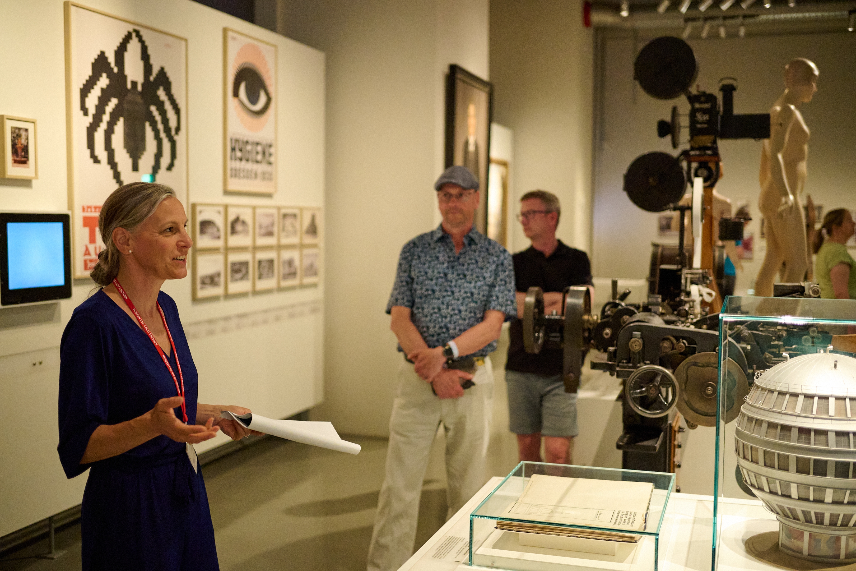 KI generiert: Das Bild zeigt eine Frau, die in einem Ausstellungsraum mit verschiedenen Exponaten spricht, darunter Poster und mechanische Geräte. Im Hintergrund stehen mehrere Personen, die der Präsentation aufmerksam folgen.