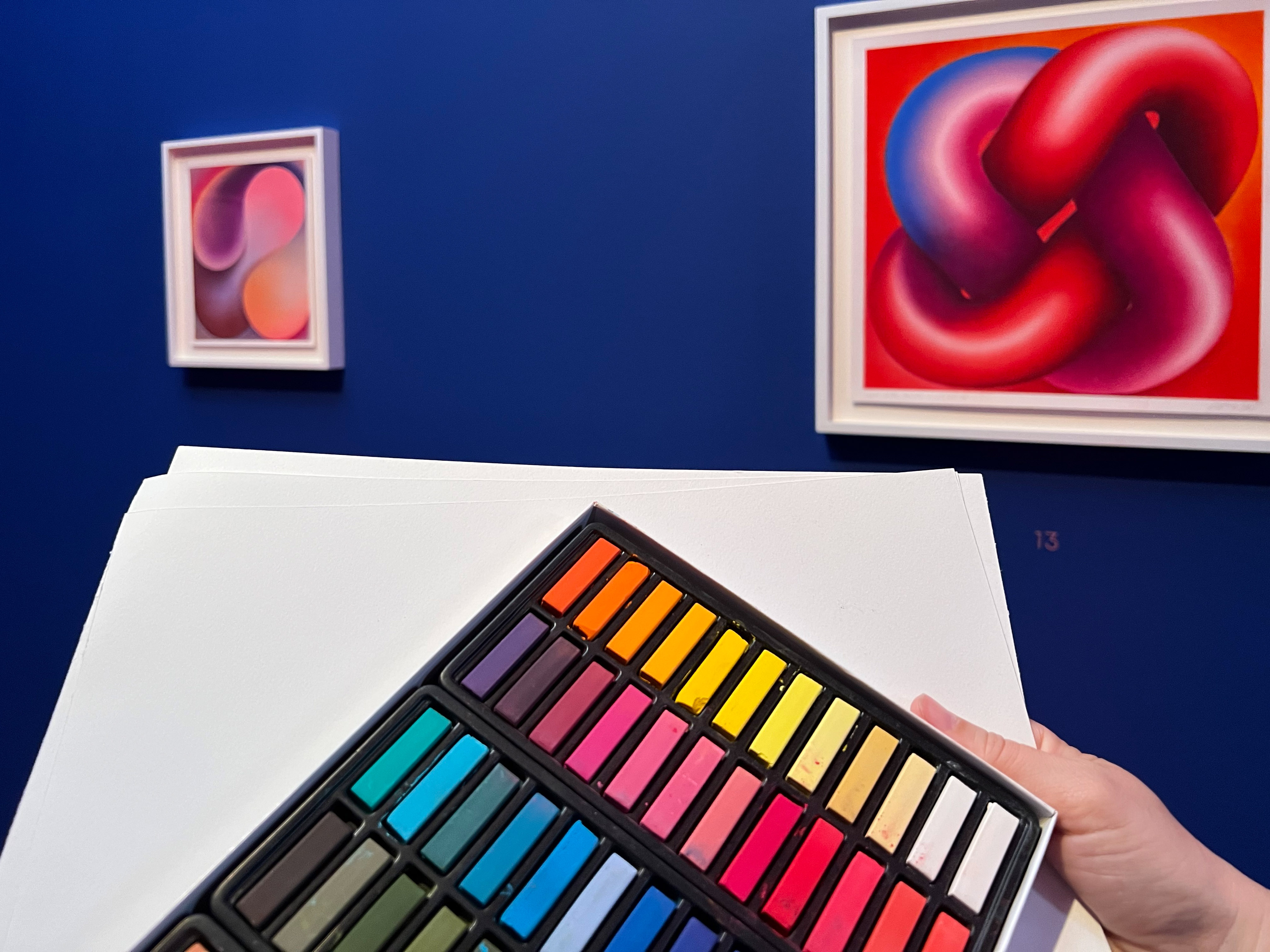 KI generiert: Das Bild zeigt ein Set bunter Pastellkreiden in der Hand einer Person vor einer blauen Wand, an der zwei abstrakte Kunstwerke hängen. Der Hauptinhalt ist die Verbindung zwischen den Farben der Kreiden und der Kunstwerke im Hintergrund.