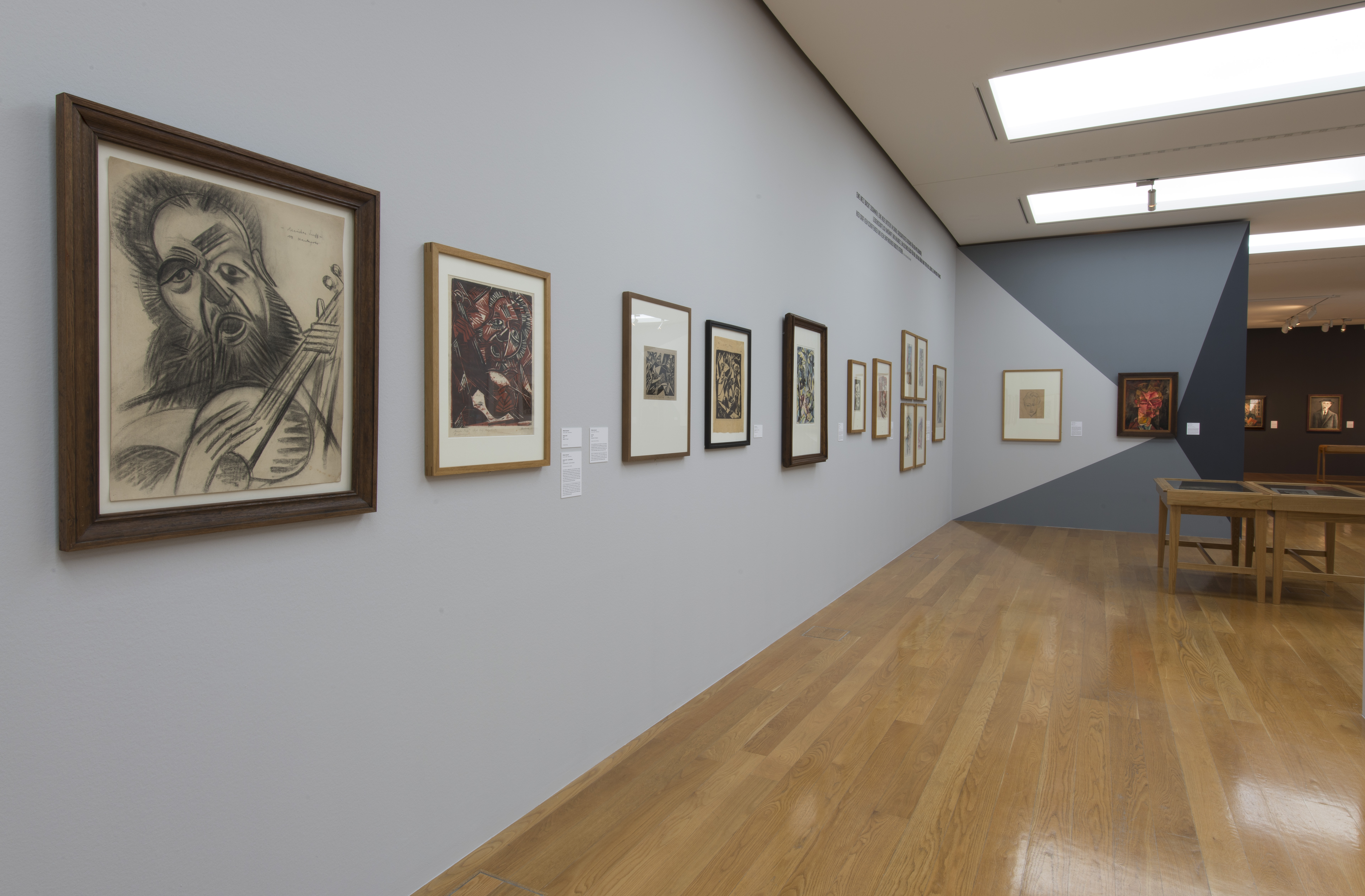 KI generiert: Das Bild zeigt eine Kunstgalerie mit mehreren gerahmten Bildern, die an einer hellgrauen Wand hängen. Der Raum hat eine moderne und minimalistische Einrichtung mit Holzboden und Tisch im Hintergrund.