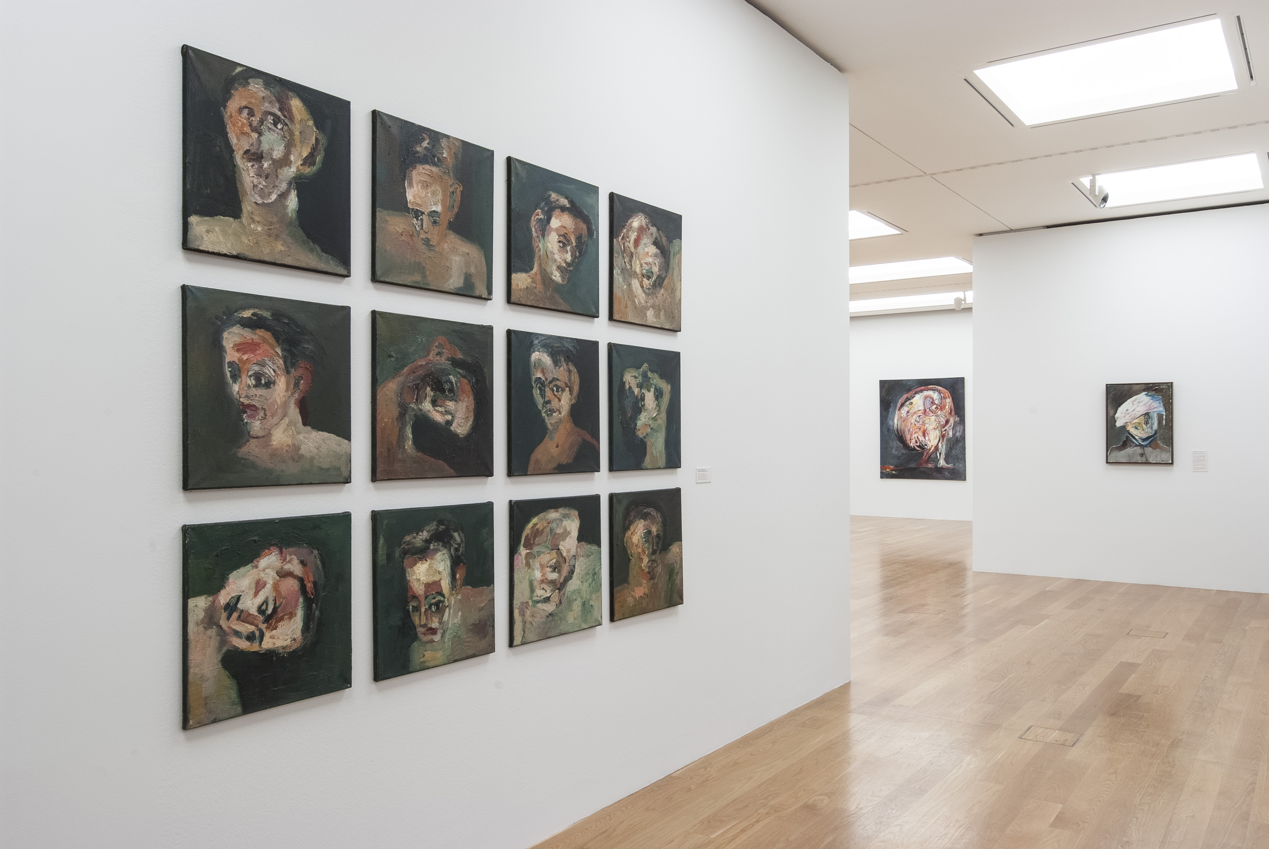 KI generiert: Das Bild zeigt eine Kunstgalerie mit mehreren expressionistischen Porträts an weißen Wänden. Der Hauptfokus liegt auf einer Serie von zwölf Gemälden, die in einem Raster angeordnet sind und verschiedene Gesichtsdetails darstellen.