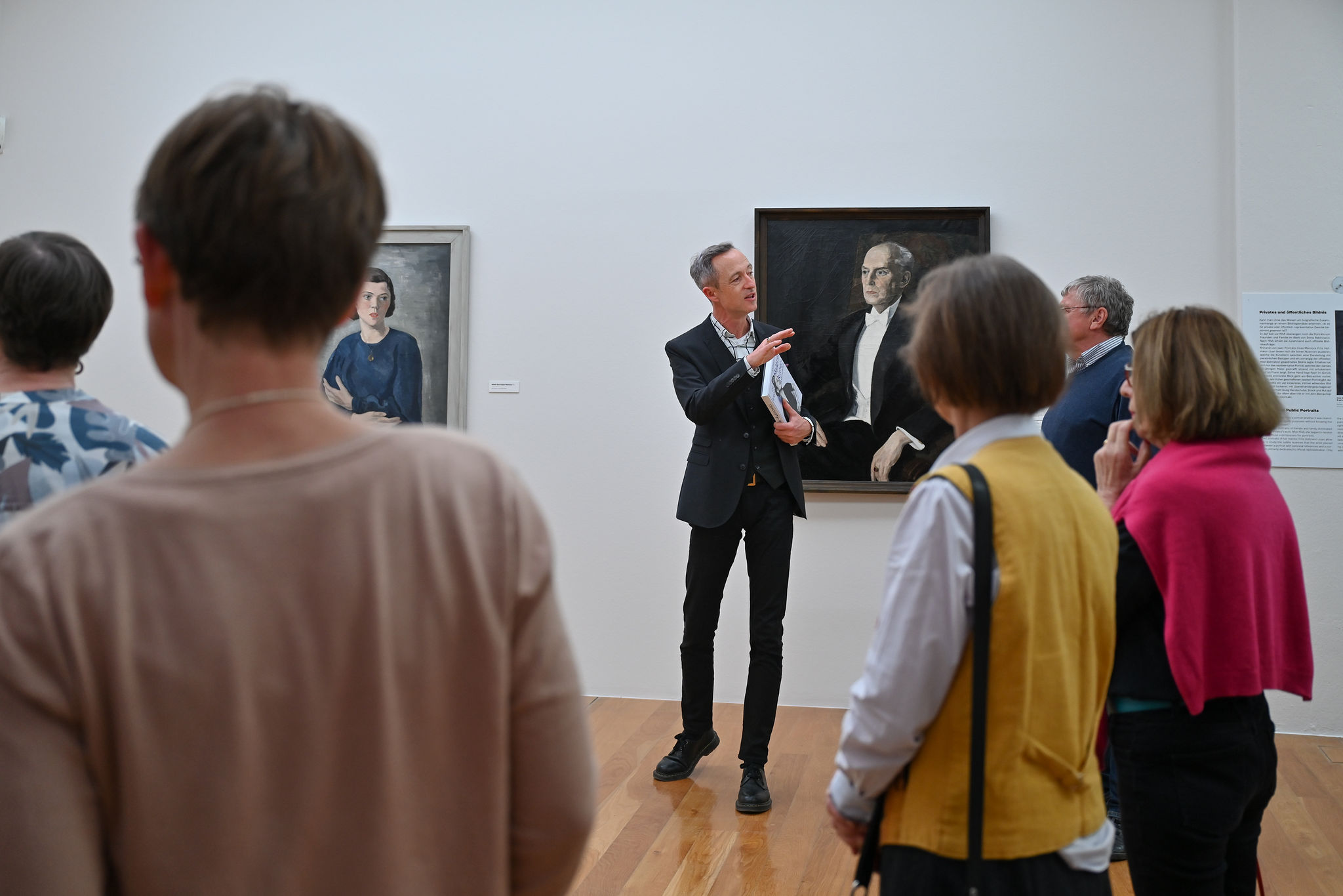 KI generiert: Das Bild zeigt eine Gruppe von Menschen, die einer Führung in einem Kunstmuseum zuhören. Im Hintergrund sind zwei Porträtgemälde zu sehen.