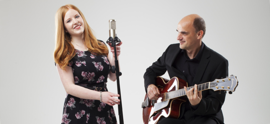 KI generiert: Das Bild zeigt eine Frau, die in ein Mikrofon singt, während ein Mann daneben Gitarre spielt. Der Hauptinhalt des Bildes ist ein musikalisches Duo.