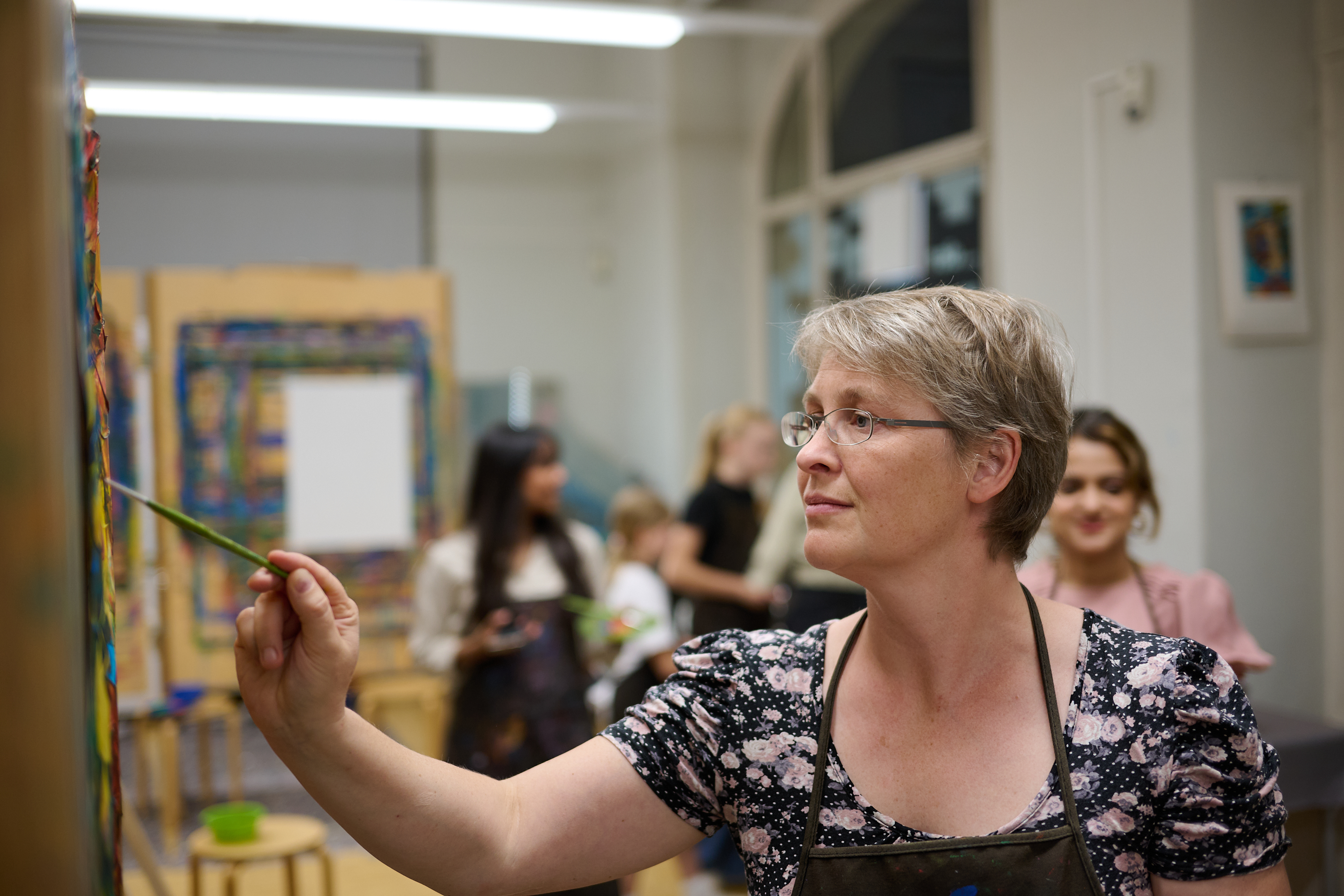 AI generated: The image shows a woman painting a work of art while other people take part in a creative event in the background. The focus is on the woman's concentrated activity with the paintbrush.