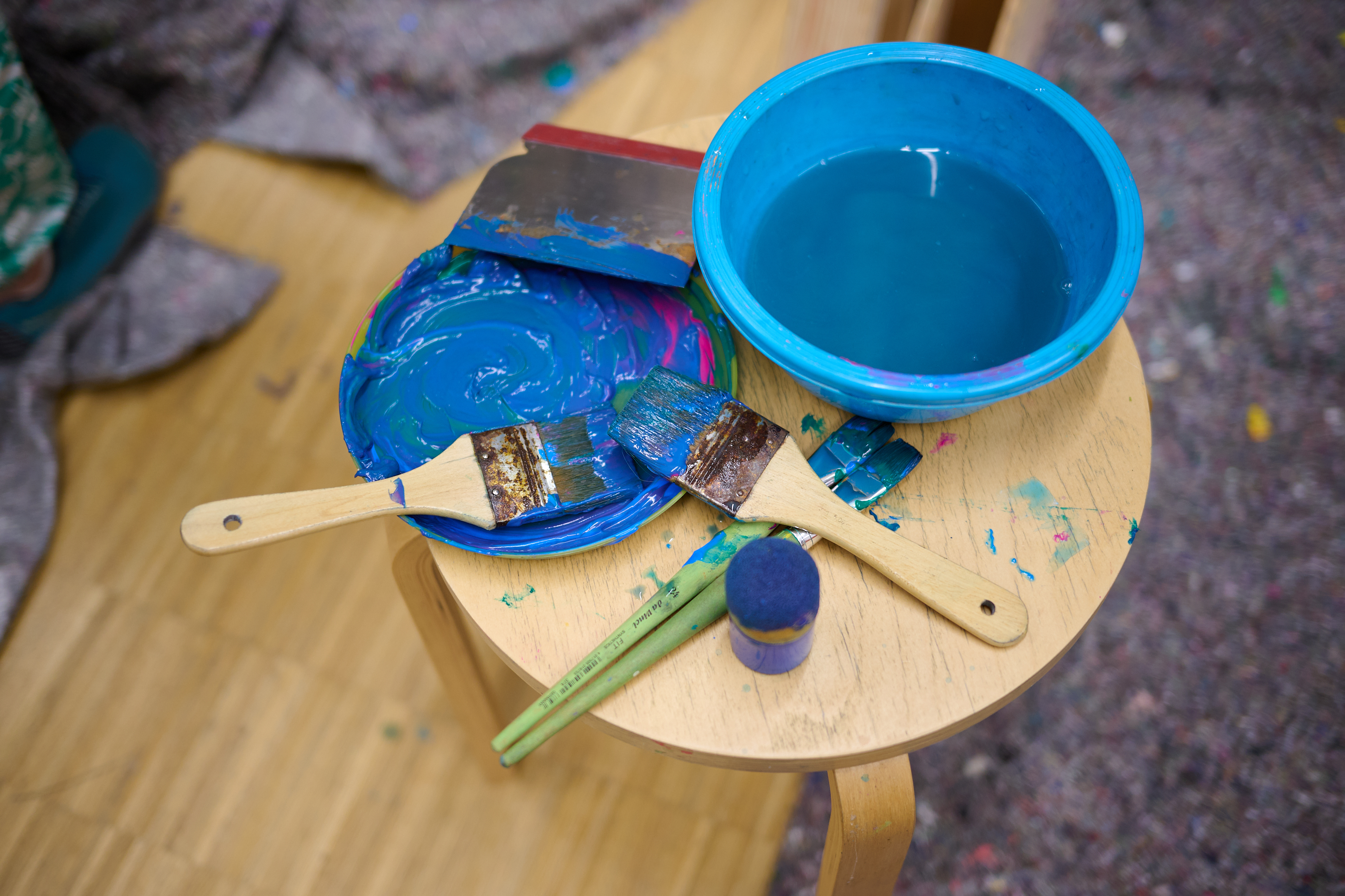 KI generiert: Auf dem Bild sind Malutensilien auf einem Hocker zu sehen, darunter Pinsel, ein Palettmesser und ein runder Behälter mit blauer Farbe. Daneben steht eine Schüssel mit Wasser.