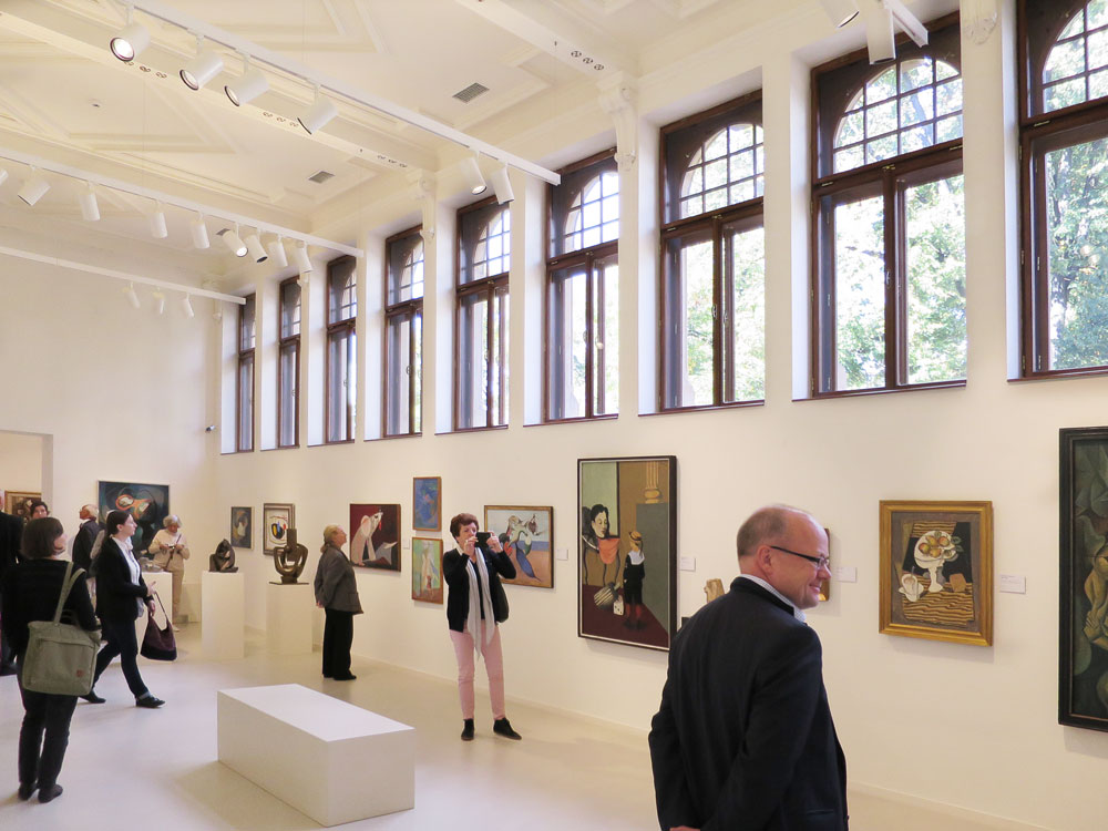 KI generiert: Das Bild zeigt eine Kunstgalerie, in der mehrere Personen Kunstwerke betrachten. Die Galerie hat hohe Fenster und Wände, an denen Gemälde hängen, und es gibt auch einige Skulpturen.