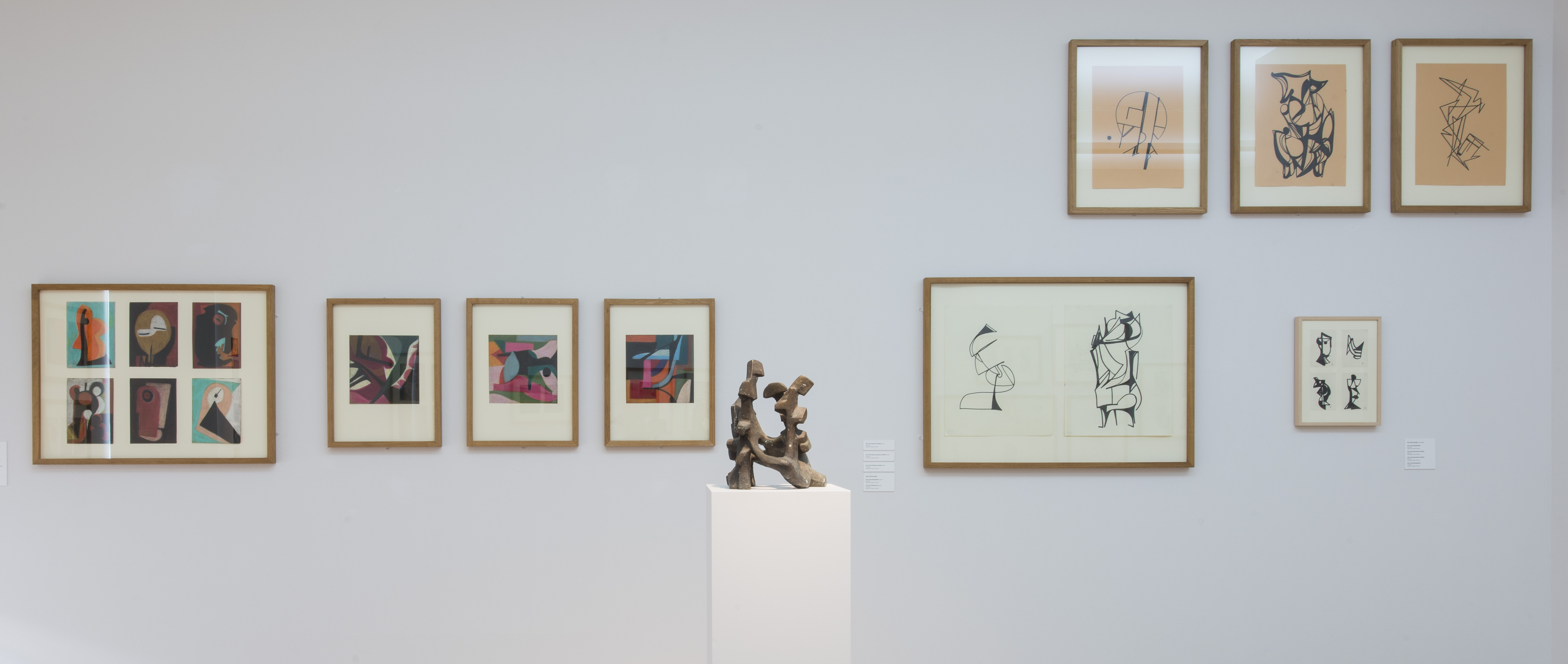 KI generiert: Das Bild zeigt eine Ausstellung von Kunstwerken an einer weißen Wand, bestehend aus Zeichnungen in Rahmen und einer Skulptur in der Mitte auf einem Podest. Der Hauptinhalt ist die Präsentation moderner Kunst in einem Galerieumfeld.
