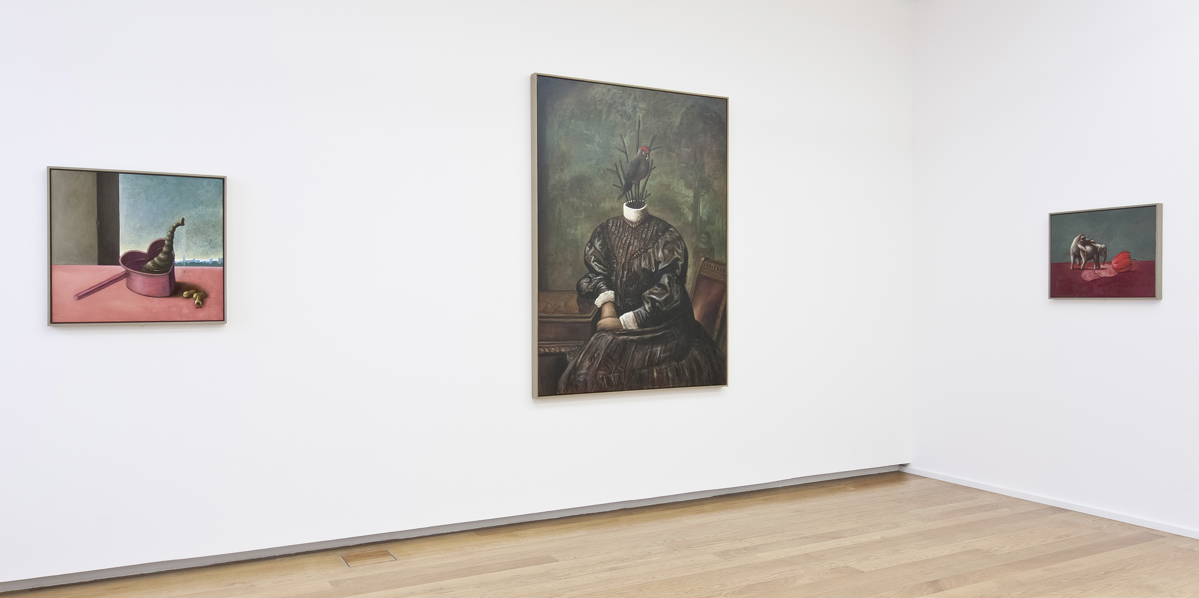 KI generiert: Das Bild zeigt eine Kunstgalerie mit drei surrealistischen Gemälden an der Wand. Die Werke enthalten ungewöhnliche Elemente, wie ein Porträt einer Person mit einem Vogelkopf und Stillleben mit teils surrealen Motiven.