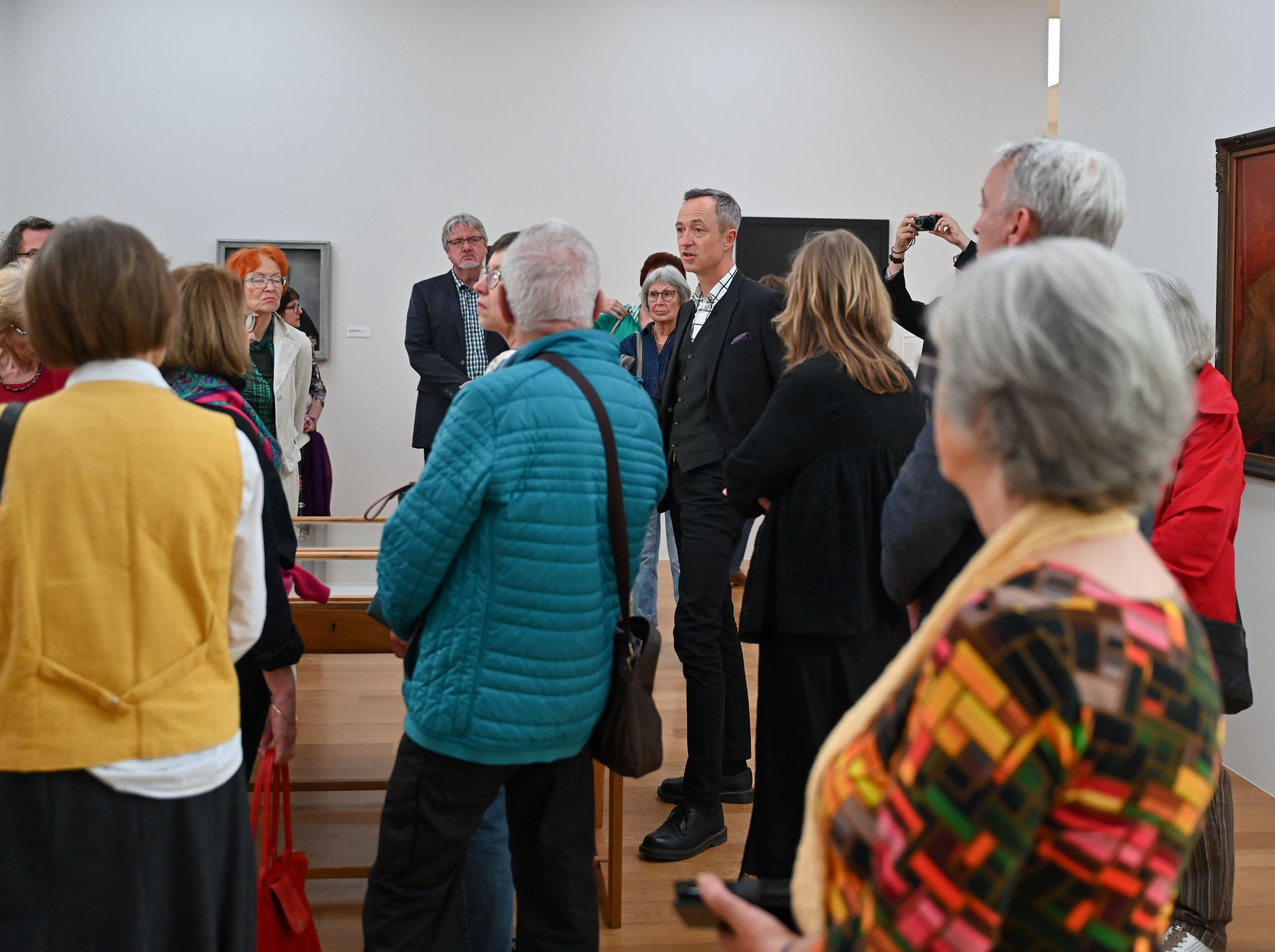 KI generiert: Das Bild zeigt eine Gruppe von Menschen in einer Galerie oder einem Museum, die anscheinend einer Führung lauschen. Im Hintergrund sind mehrere Gemälde zu sehen.