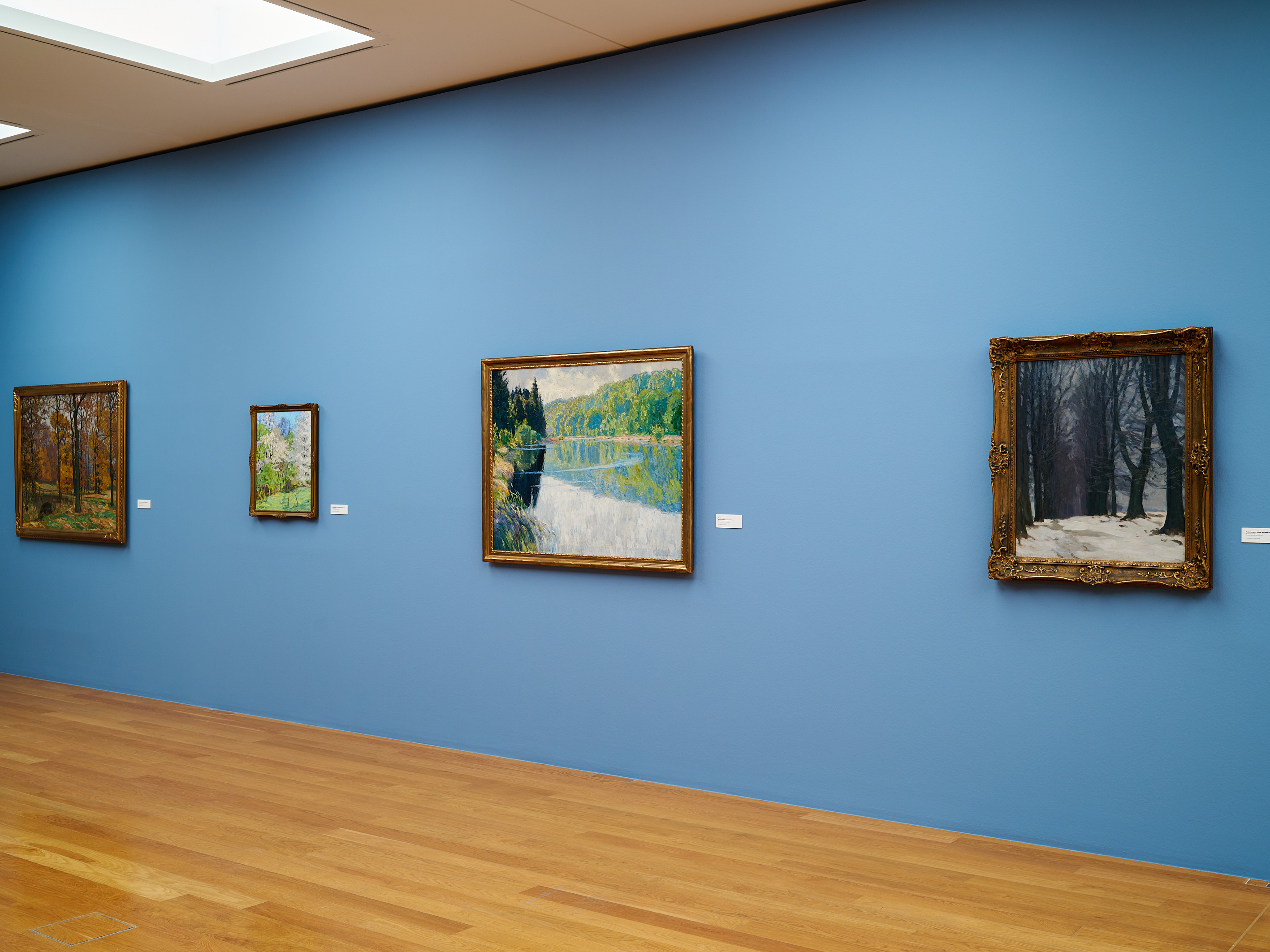 KI generiert: Das Bild zeigt eine Galerie mit einer blauen Wand, an der vier gerahmte Bilder hängen, die Landschaften darstellen. Der Raum ist mit Holzfußboden und einer modernen Deckenbeleuchtung ausgestattet.