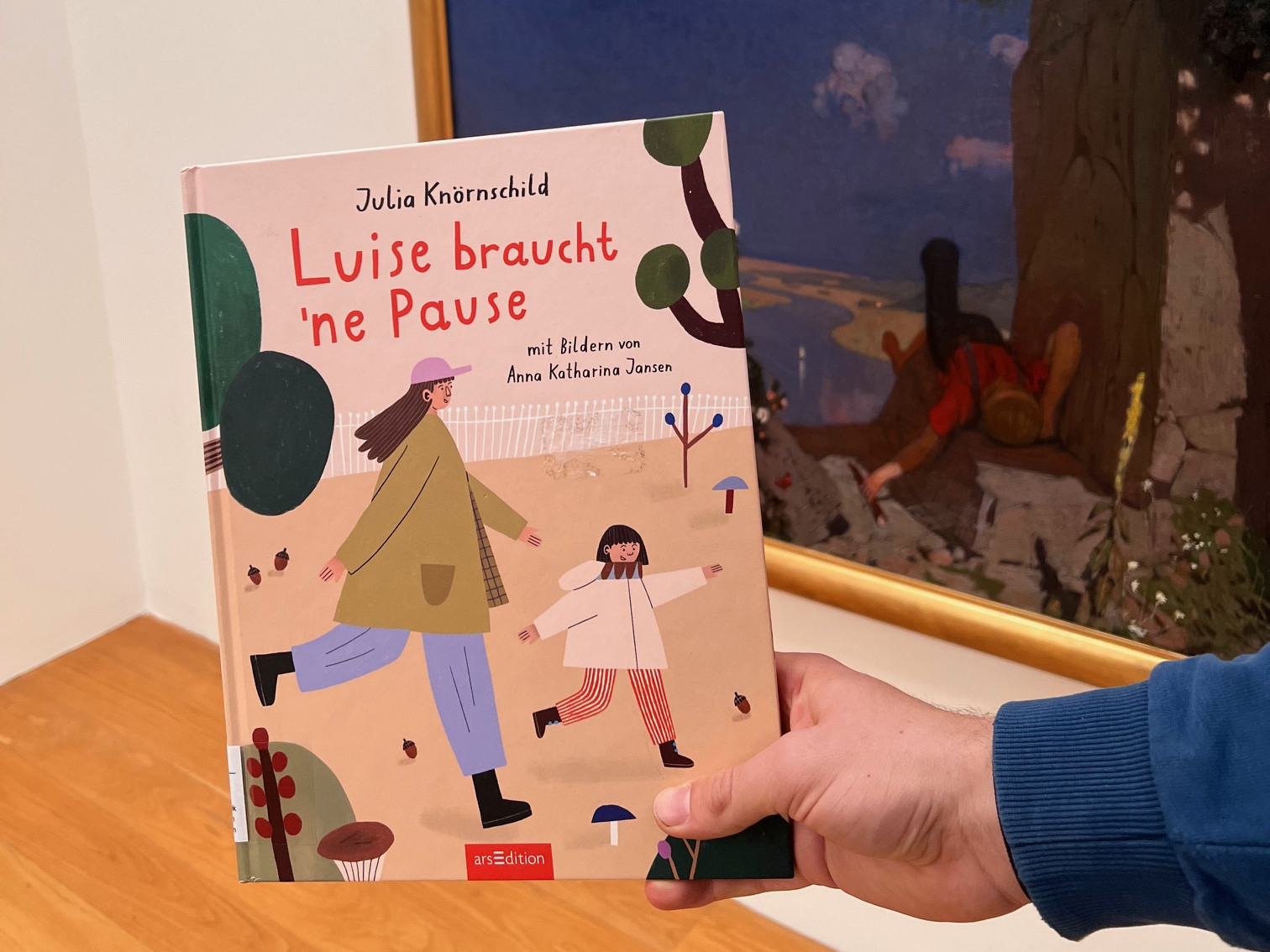 KI generiert: Das Bild zeigt eine Person, die ein Kinderbuch mit dem Titel "Luise braucht 'ne Pause" von Julia Knobrnshild hält. Im Hintergrund ist ein Gemälde mit einer natürlichen Landschaft zu sehen.