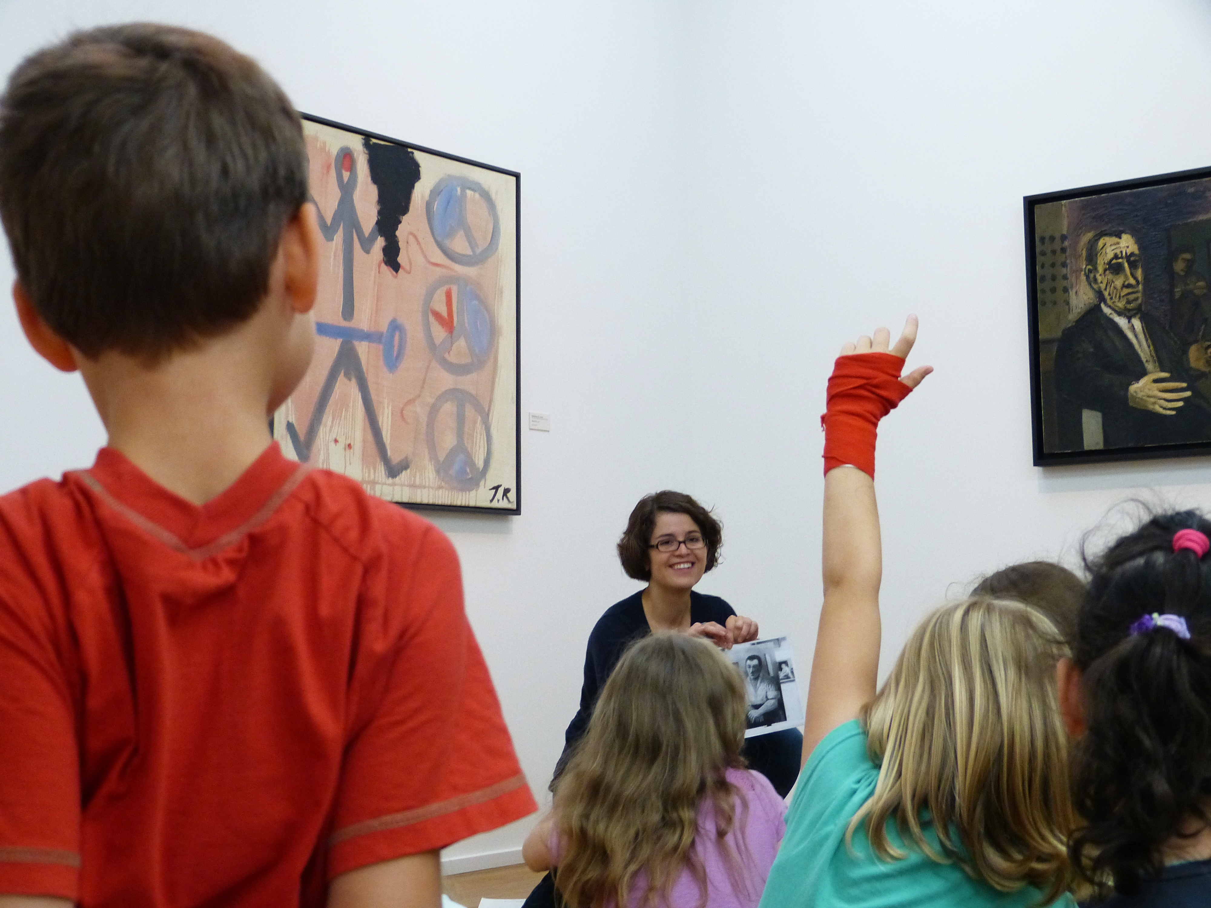 KI generiert: Das Bild zeigt eine Gruppe von Kindern in einem Kunstmuseum, die an einer Führung oder einem Workshop teilnehmen. Eine lächelnde Frau sitzt vor den Kindern und hält ein Bild hoch, während ein Kind die Hand hebt, um eine Frage zu stellen.