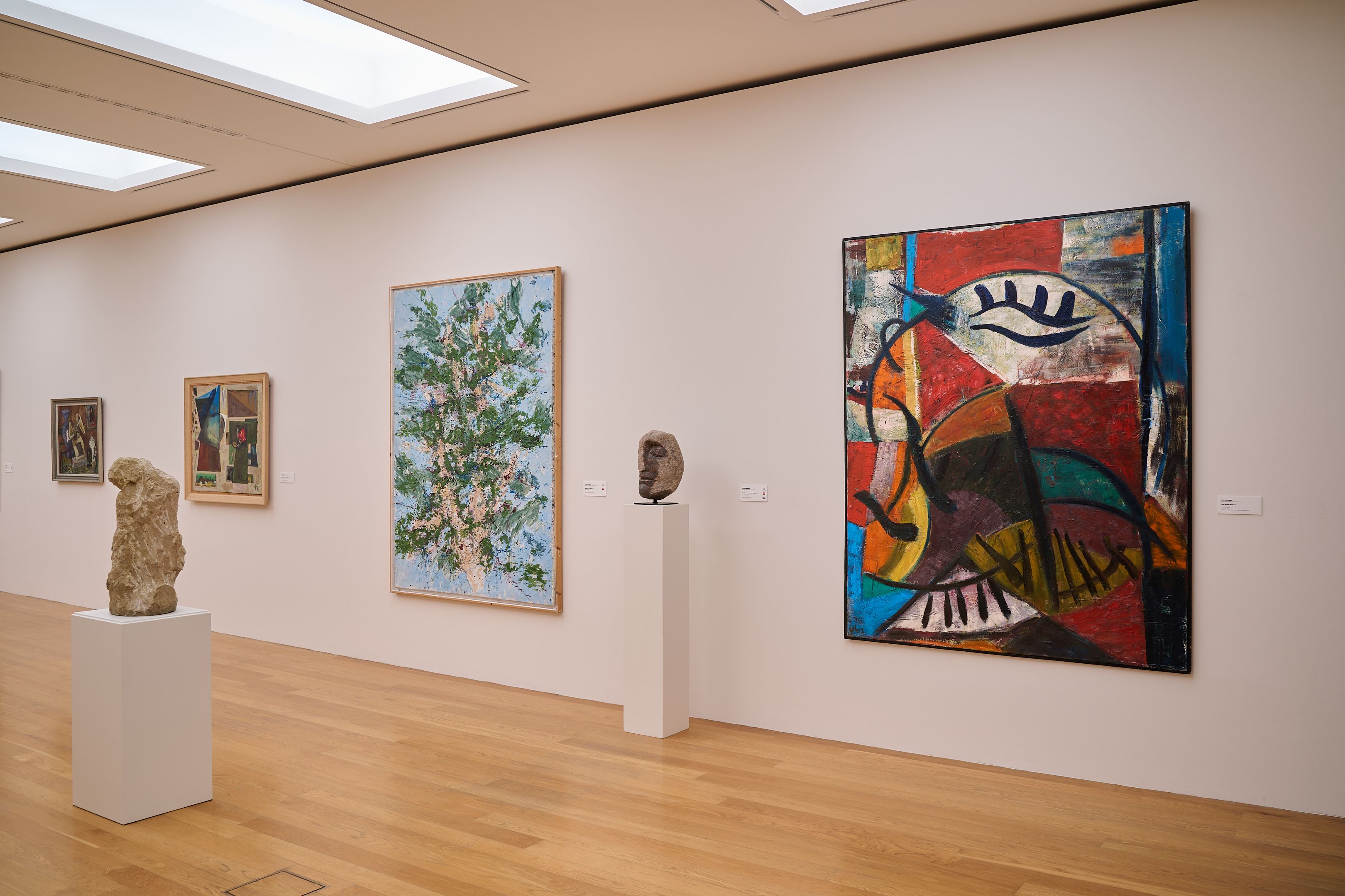 KI generiert: Das Bild zeigt eine Galerie mit verschiedenen Kunstwerken, darunter Gemälde und Skulpturen, die an einer weißen Wand ausgestellt sind. Der Raum hat einen Holzfußboden und eine helle, moderne Beleuchtung.