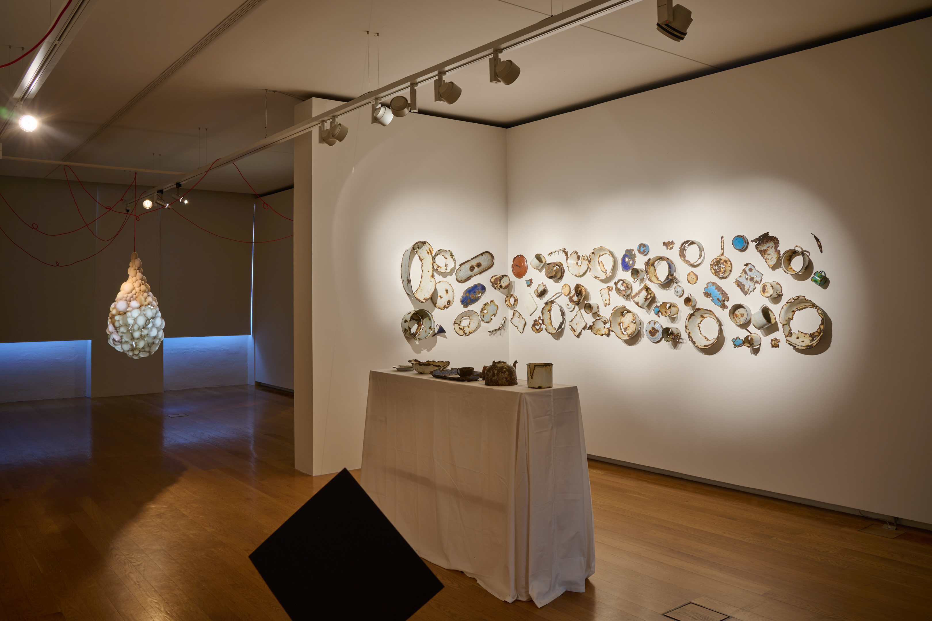 AI generated: The image shows a modern art exhibition in a room with illuminated objects on the wall and a sculpture-like installation on the left in the background. In the foreground is a table covered with a white cloth with other art objects.