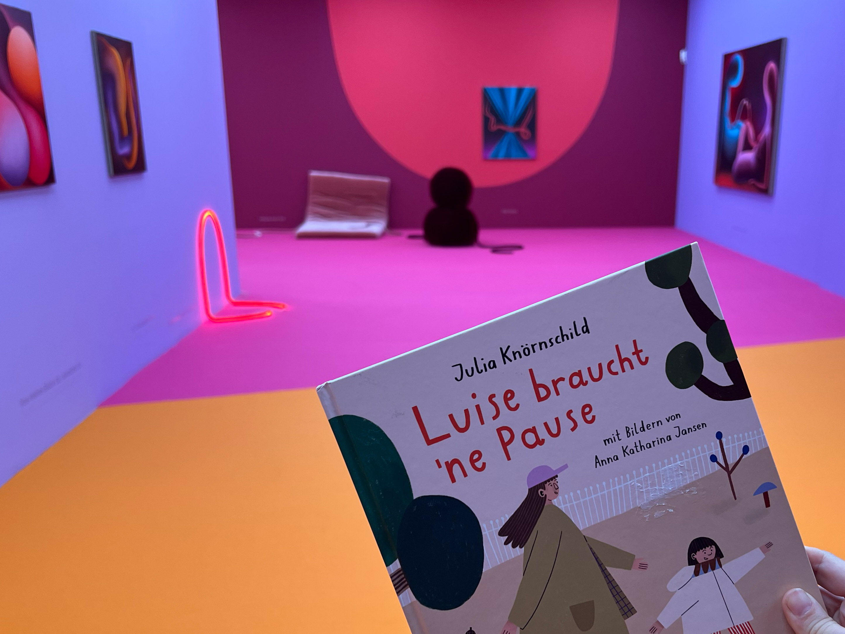 KI generiert: Das Bild zeigt das Buch "Luise braucht 'ne Pause" vor einem farbenfrohen, modernen Kunstraum mit abstrakten Gemälden und Installationen. Im Vordergrund ist das Buchcover zu erkennen, während der Raum im Hintergrund eine lebhafte Farbkombination aufweist.