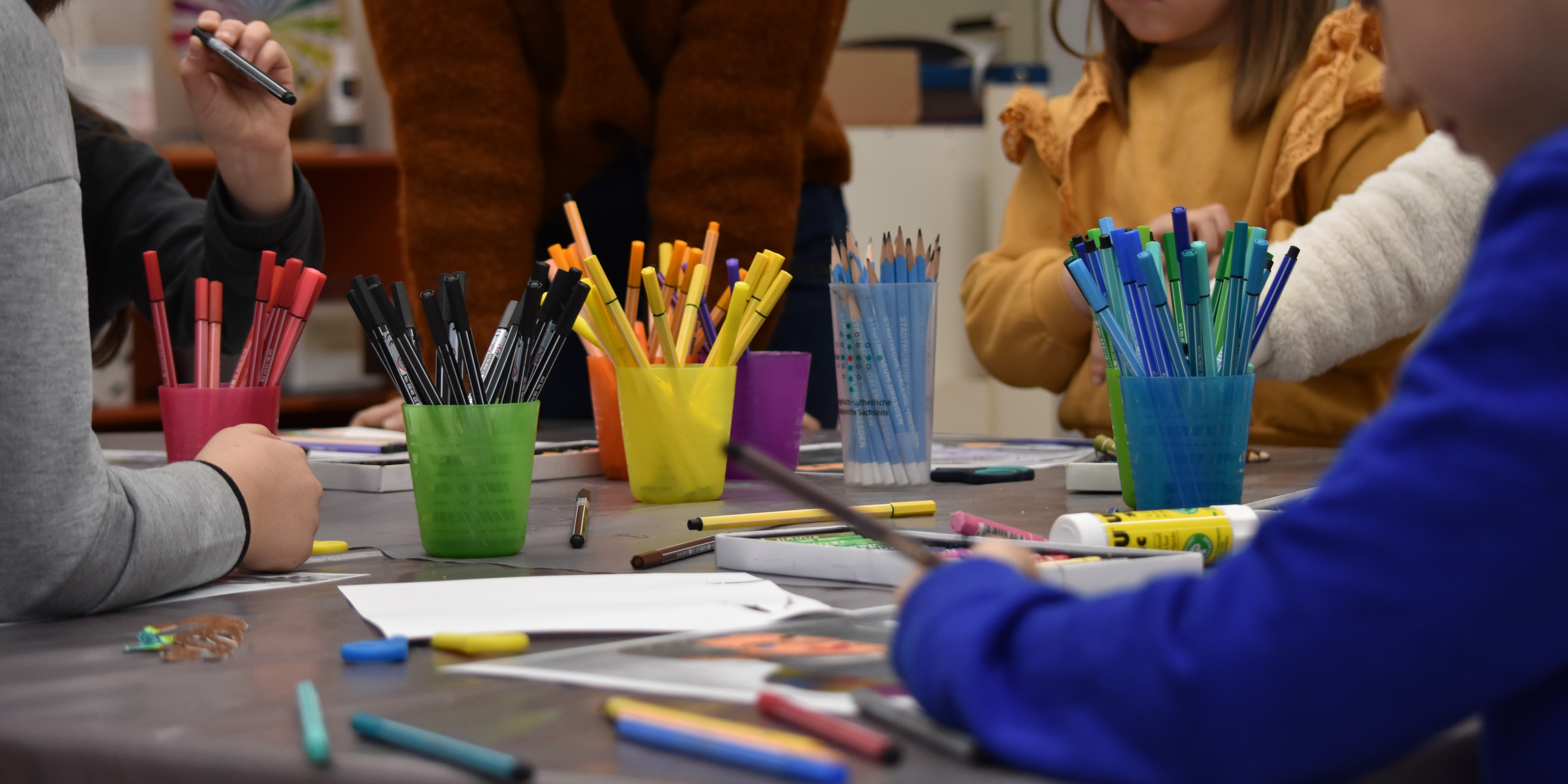 KI generiert: Das Bild zeigt einen Tisch mit bunten Bechern voller Stifte, an dem mehrere Personen gemeinsam kreativ arbeiten. Der Schwerpunkt liegt auf den verschiedenen Stiften und Händen, die sie benutzen.