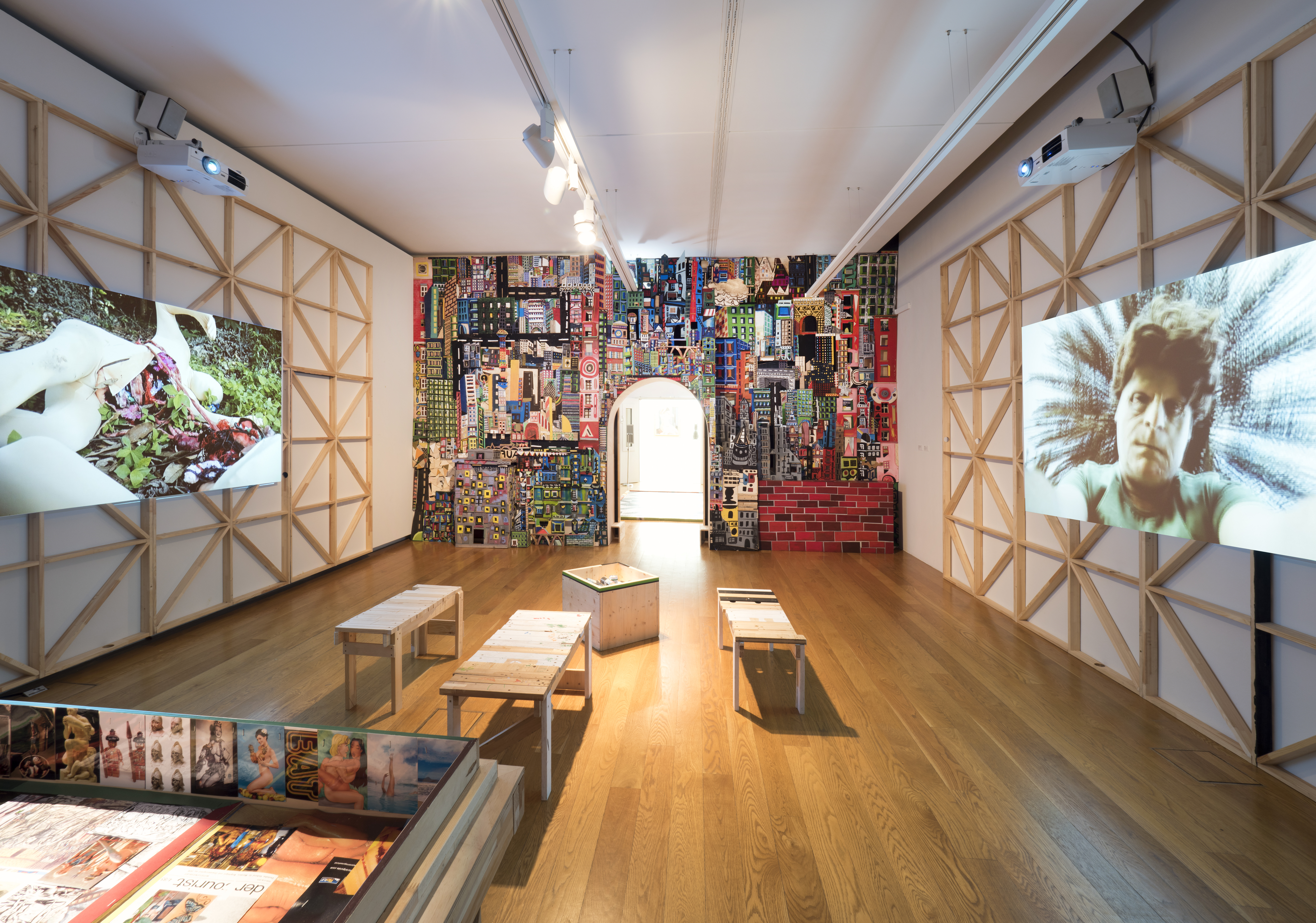 AI generated: The image shows an exhibition space with projections on the walls and an artistically designed wall of colourful billboards. There are simple wooden benches and a table in the room.